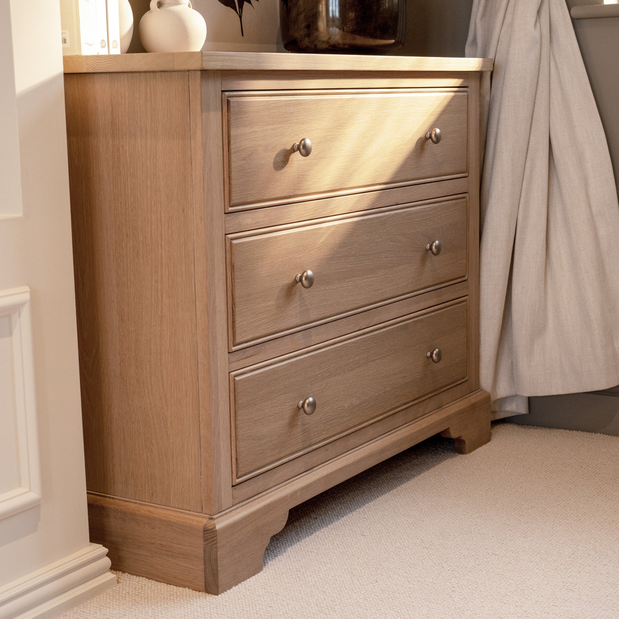 The Amberley 3 Chest of Drawers in Light Oak, with three spacious drawers and silver knobs, stands elegantly on a beige carpet in a softly lit room.