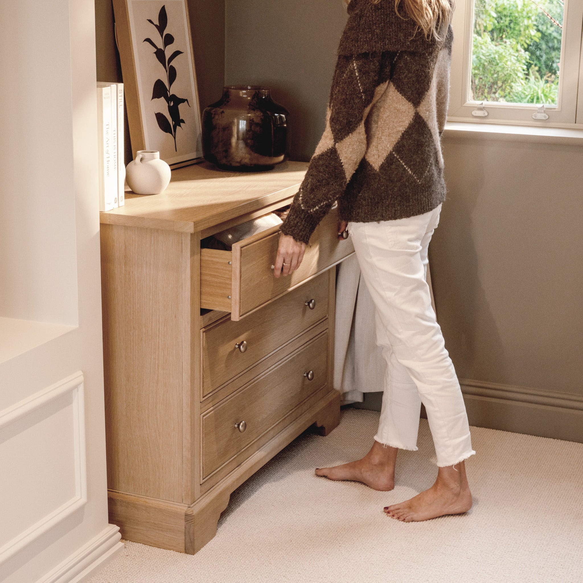 A barefoot woman in white pants opens a drawer of the Amberley 3 Chest of Drawers in Light Oak, adding warmth to a cozy, softly lit room.