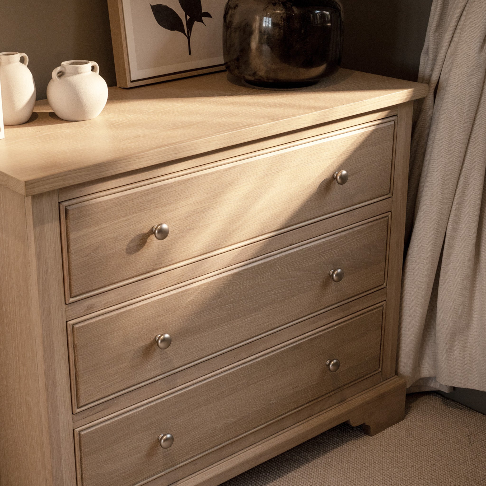 The Amberley 3 Chest of Drawers in Light Oak sits by a window, topped with ceramic vases, framed art, and a dark vase—part of the elegant Amberley collection.