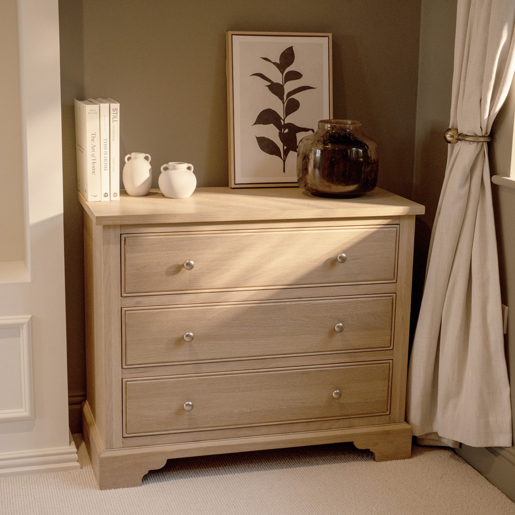 The Amberley 3 Chest of Drawers in Light Oak features three drawers and is styled with books, vases, a dark jar, and framed leaf art on top, placed near a window.