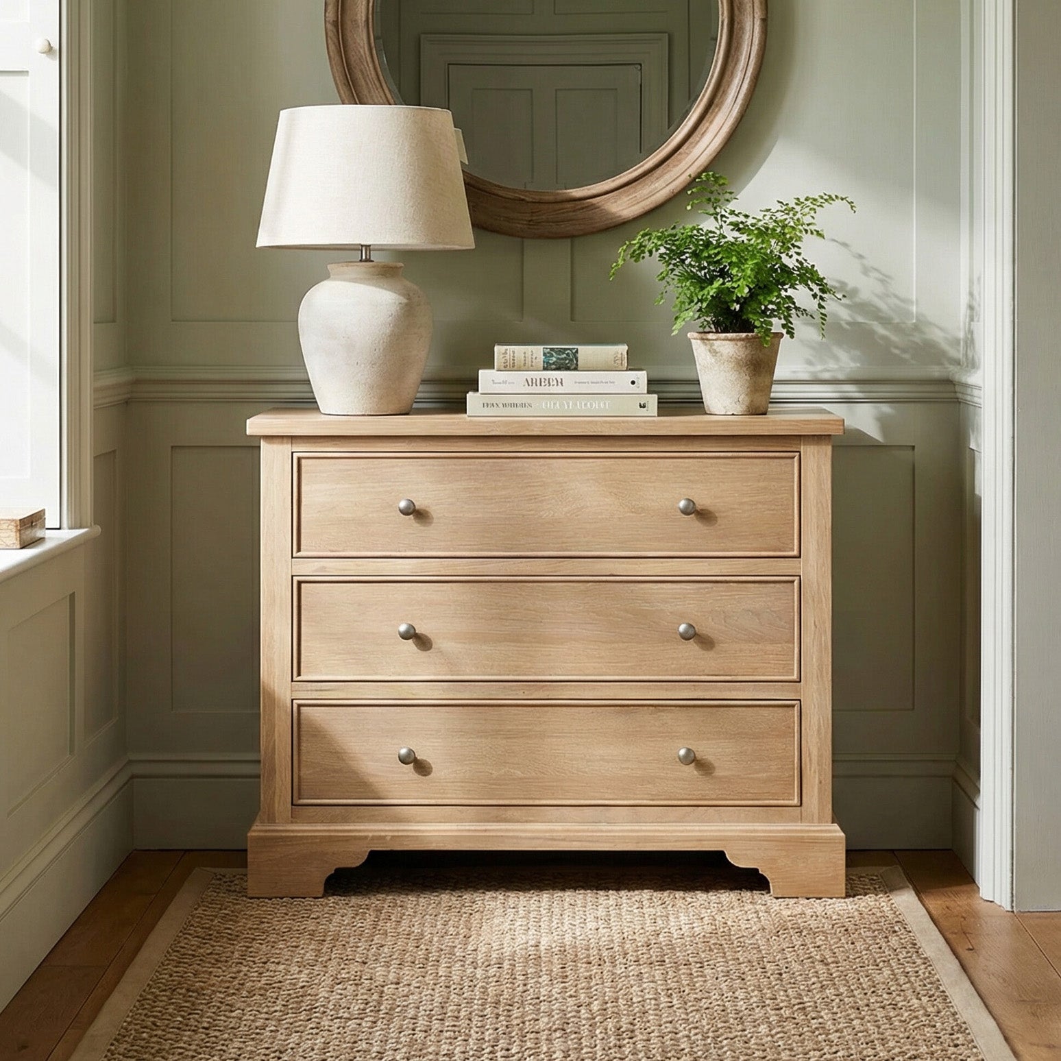 Amberley 3 Chest of Drawers in Light Oak with three drawers, topped with a lamp, books, and a potted plant, sits on a woven rug beneath a round mirror.