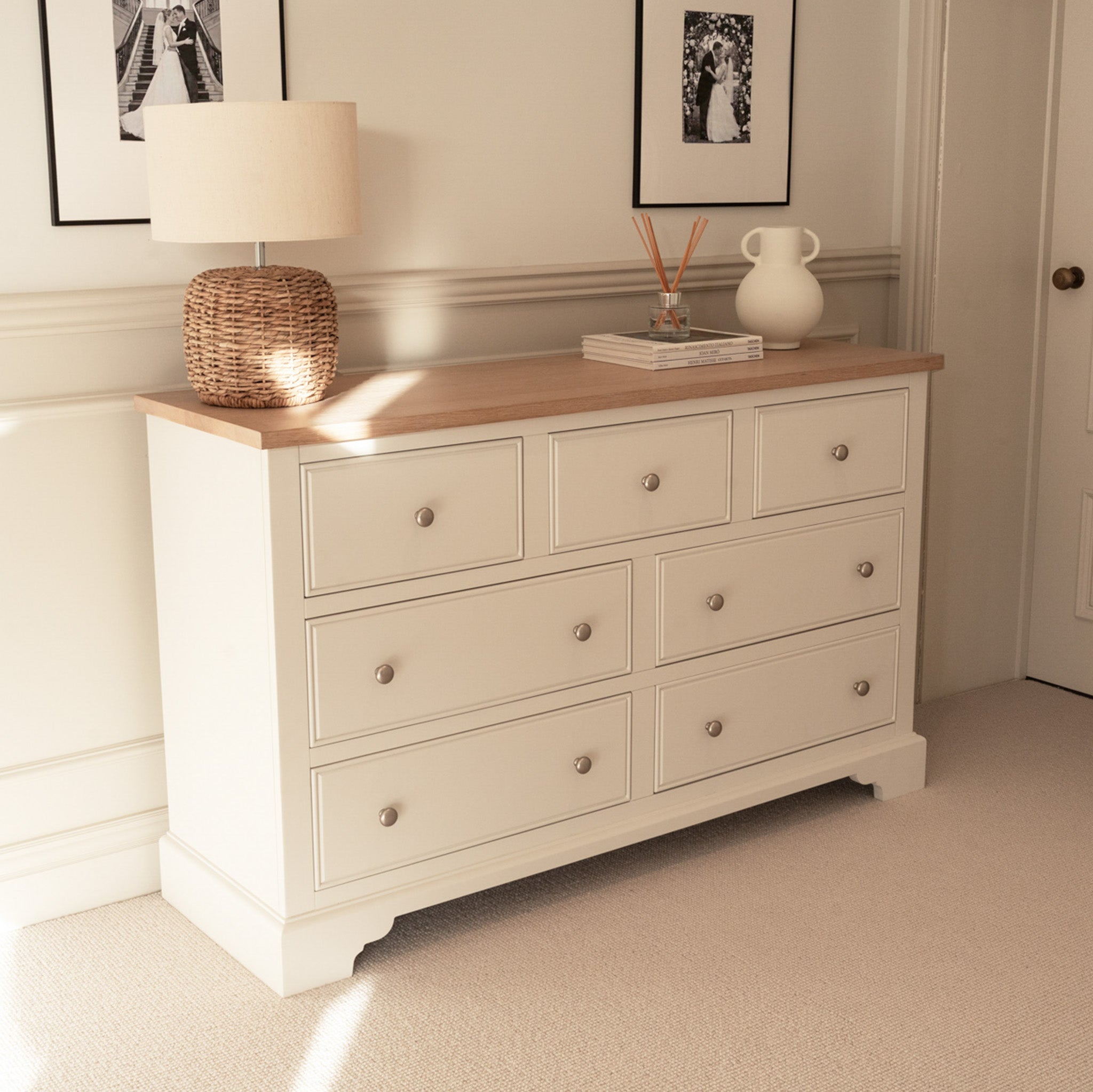 The Amberley 3 Over 4 Chest of Drawers in Warm White has an oak top and seven drawers, styled with a lamp, books, and decor above, set against a beige wall with framed photos.