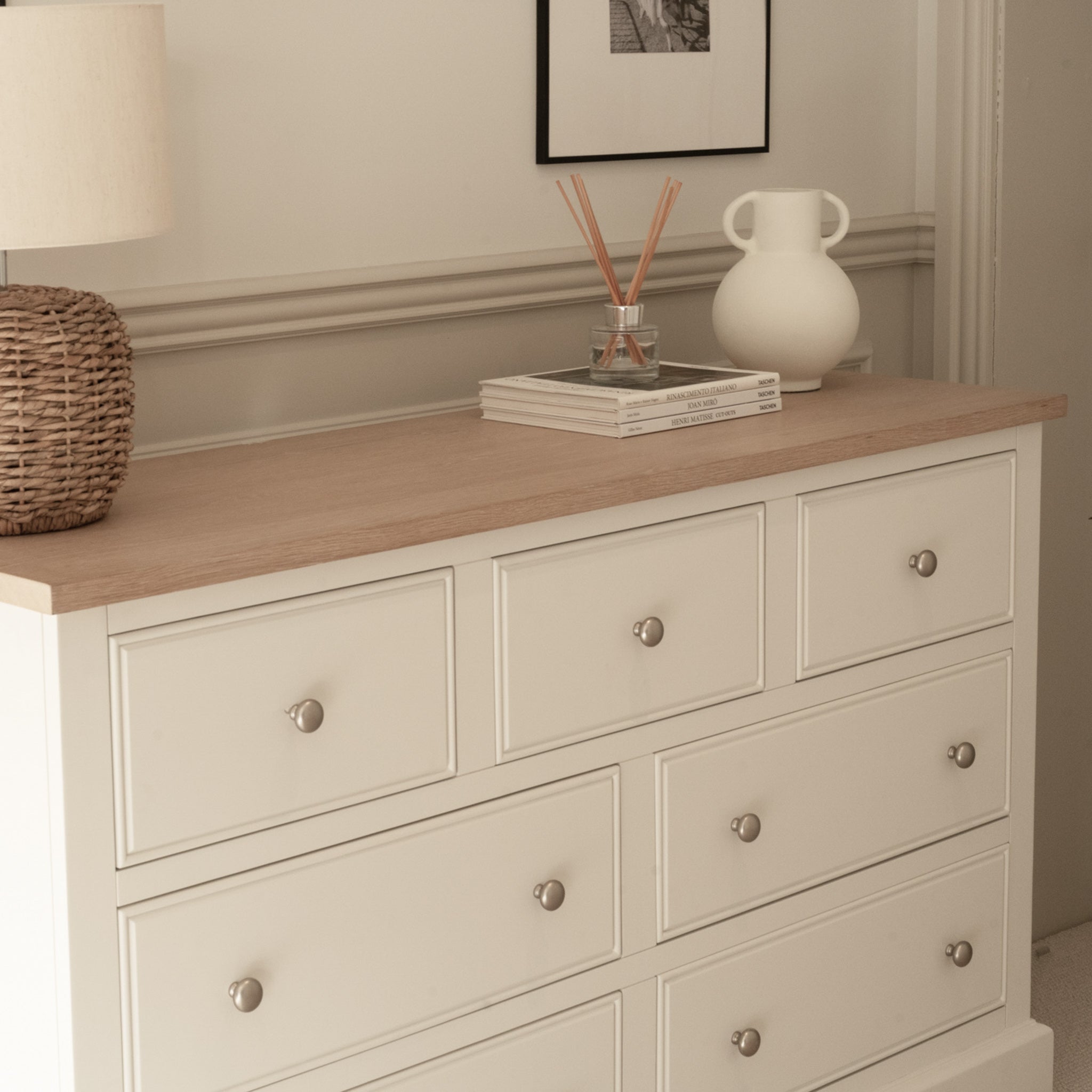 The Amberley 3 Over 4 Chest of Drawers in Warm White has an oak top, displayed with a lamp, books, and a vase.