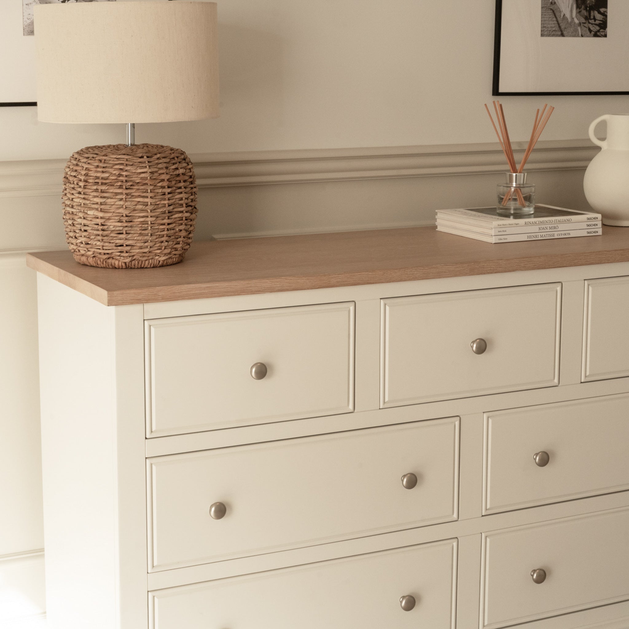 The Amberley 3 Over 4 Chest of Drawers in Warm White, accented with silver knobs, stands in a softly lit neutral room alongside a wicker lamp, books, and decorative items.