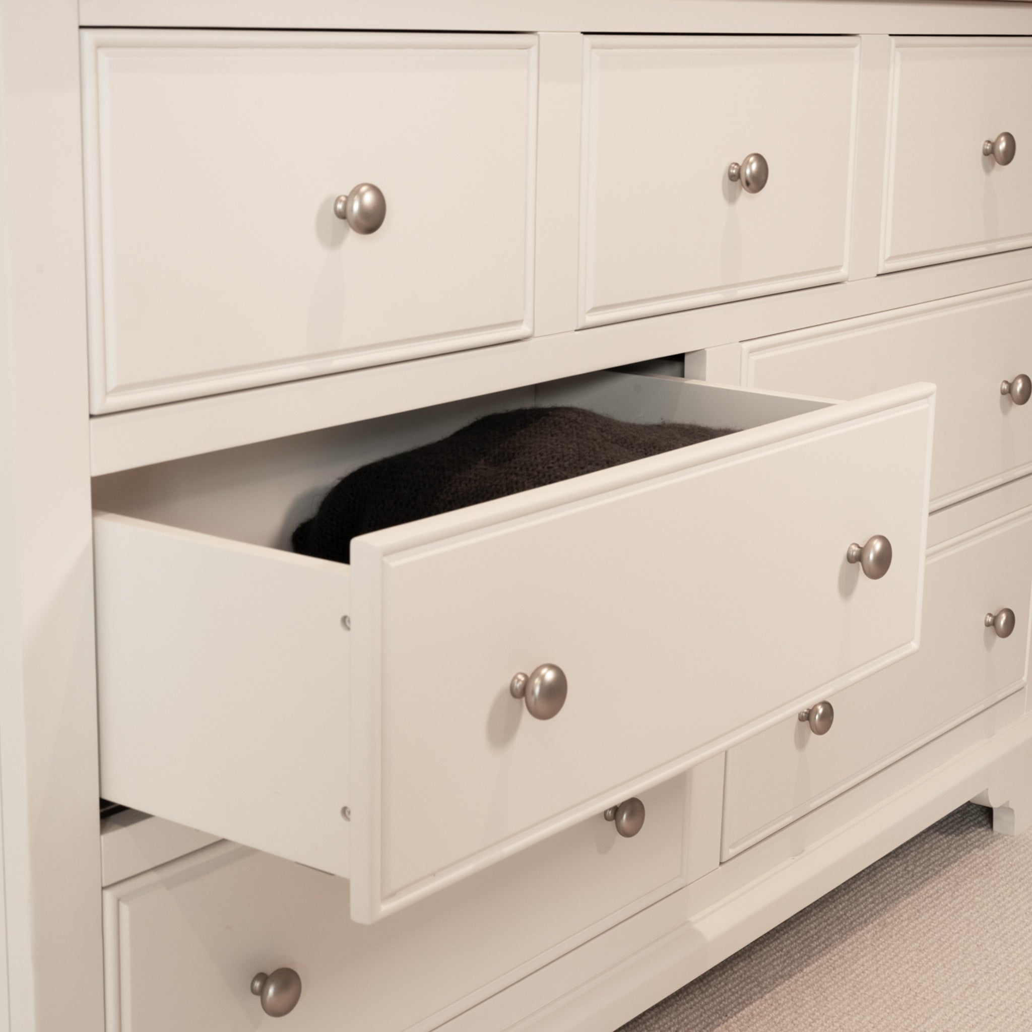 The Amberley 3 Over 4 Chest of Drawers in Warm White features an oak top and displays an open drawer with a dark garment inside.