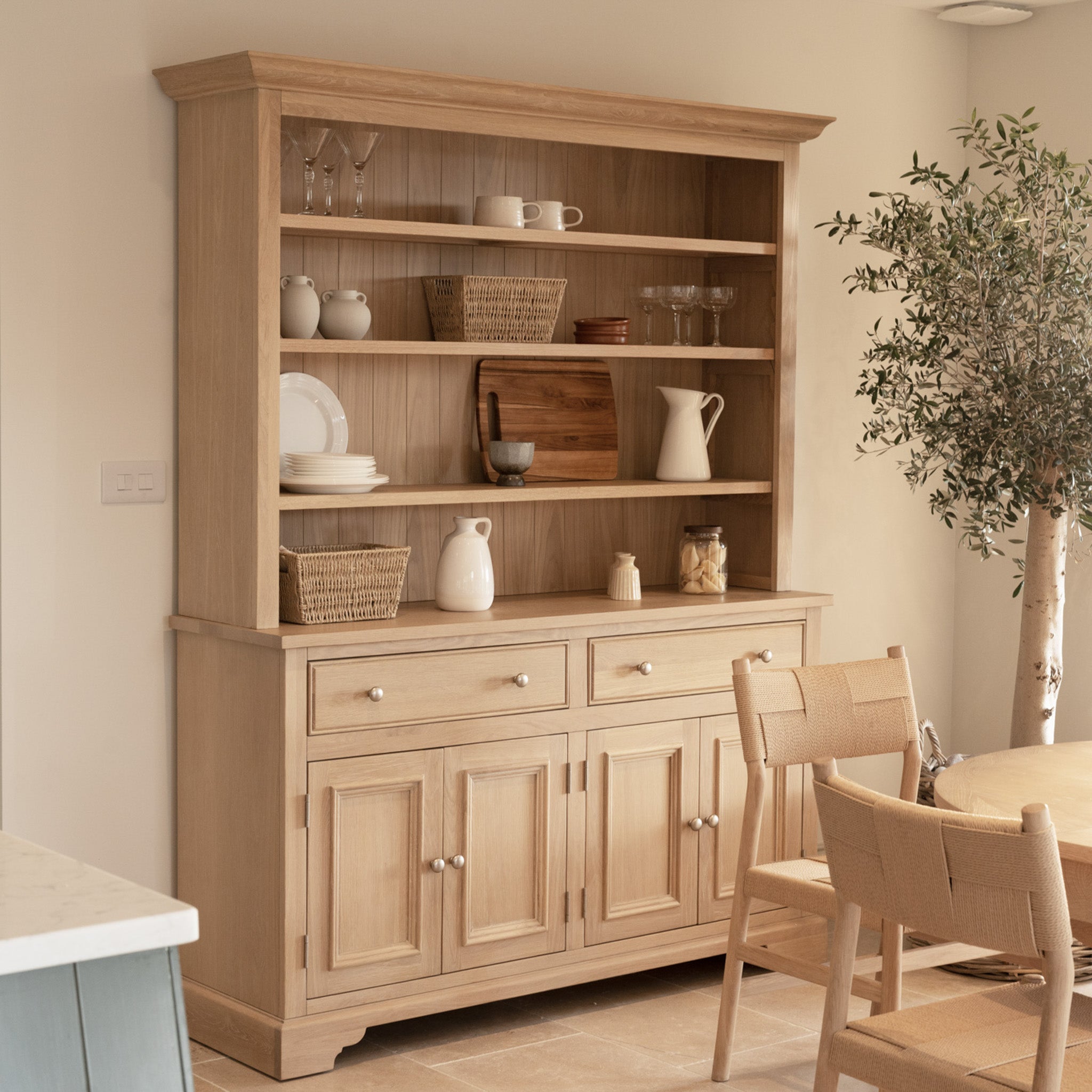 The Amberley 5ft Grand Open Dresser in Light Oak displays dishes, baskets, and glasses beside a dining table and an indoor plant.