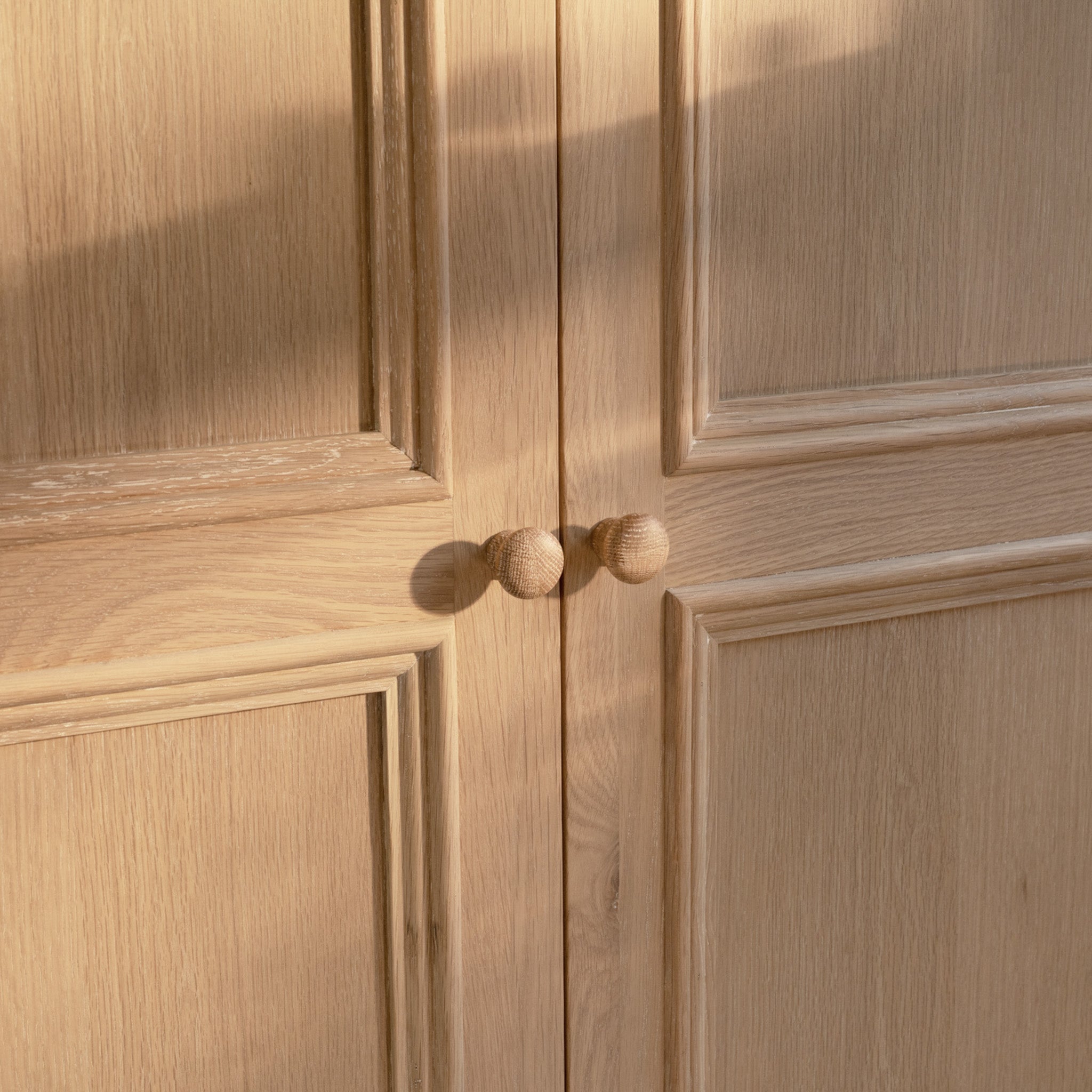 The Amberley Double Wardrobe in Light Oak features round wooden knobs and decorative panel detailing, forming part of a solid oak bedroom set and softly illuminated by sunlight.