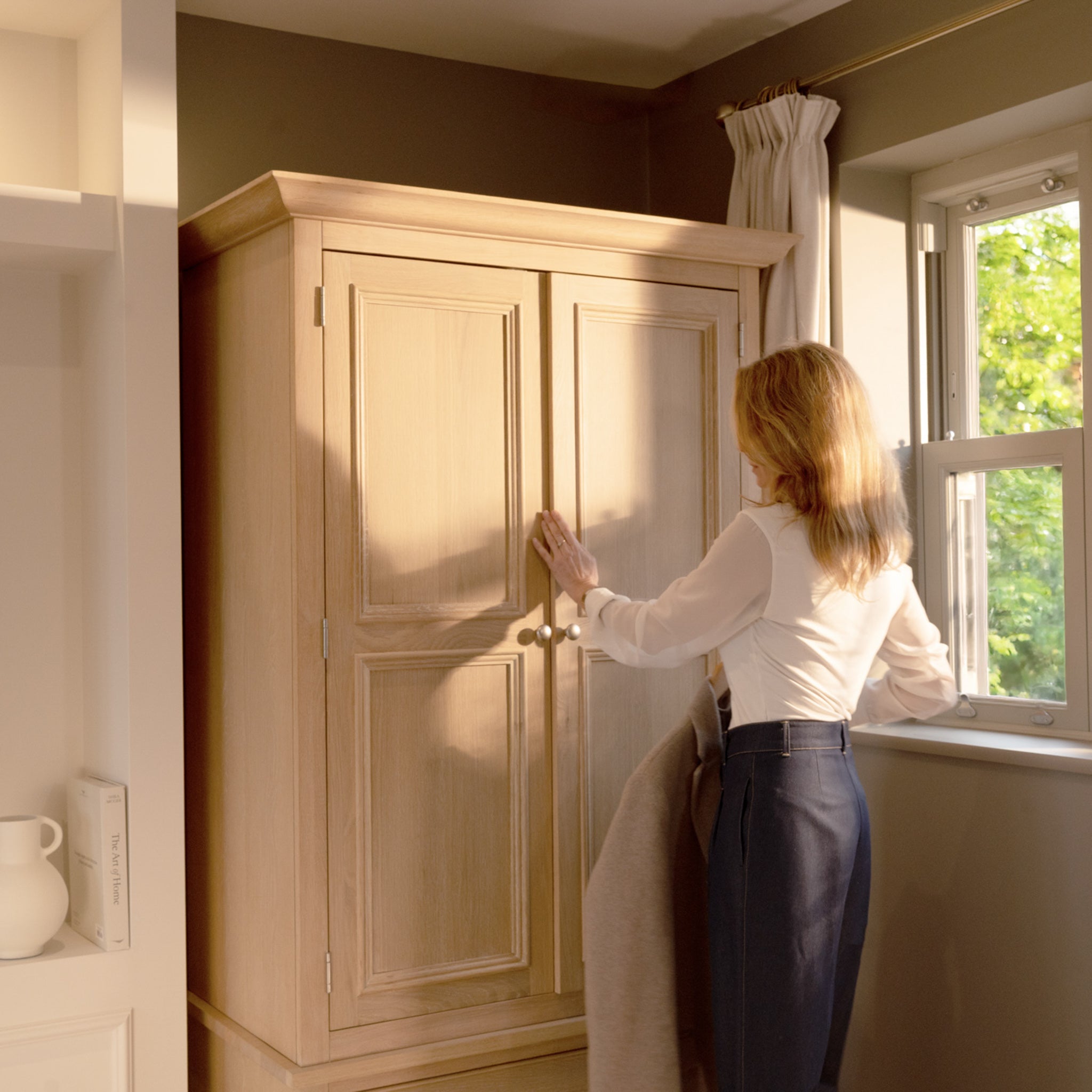 A woman in a white blouse opens the Amberley Double Wardrobe in Light Oak near a sunlit window.
