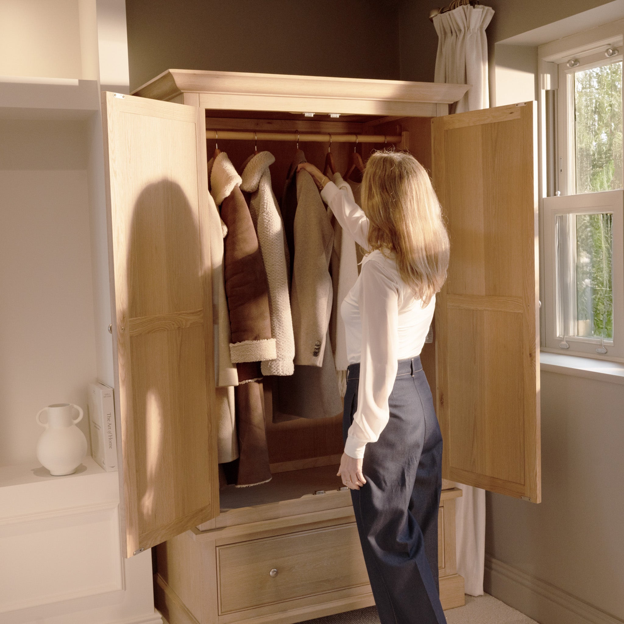 A woman reaches for a coat inside the Amberley Double Wardrobe in Light Oak, surrounded by sunlight and elegant solid oak bedroom furniture.