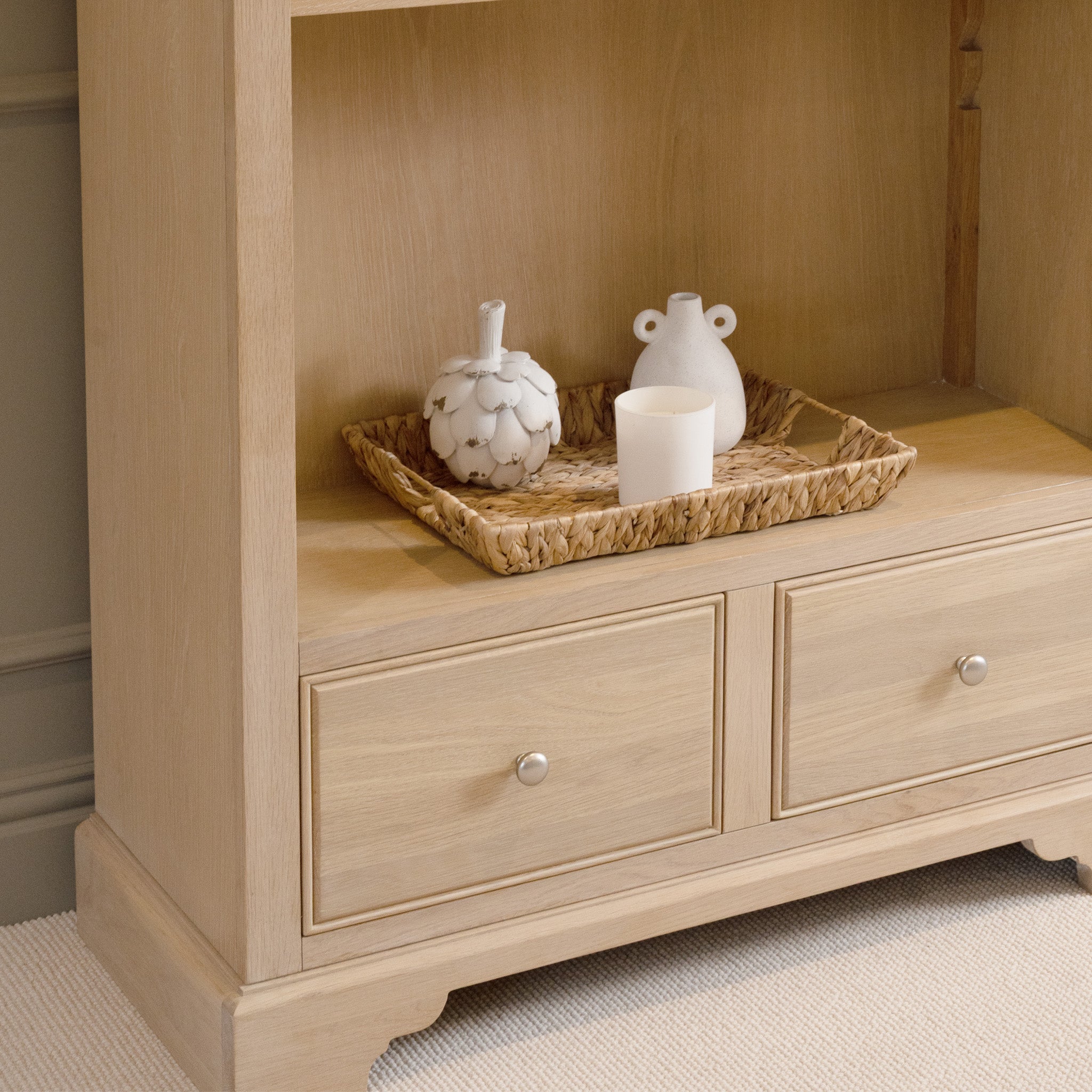 The Amberley Large Bookcase in Light Oak is styled with a wicker tray, white candle, and two decorative vases on a wooden shelf with drawers below for a cozy, modern country look.