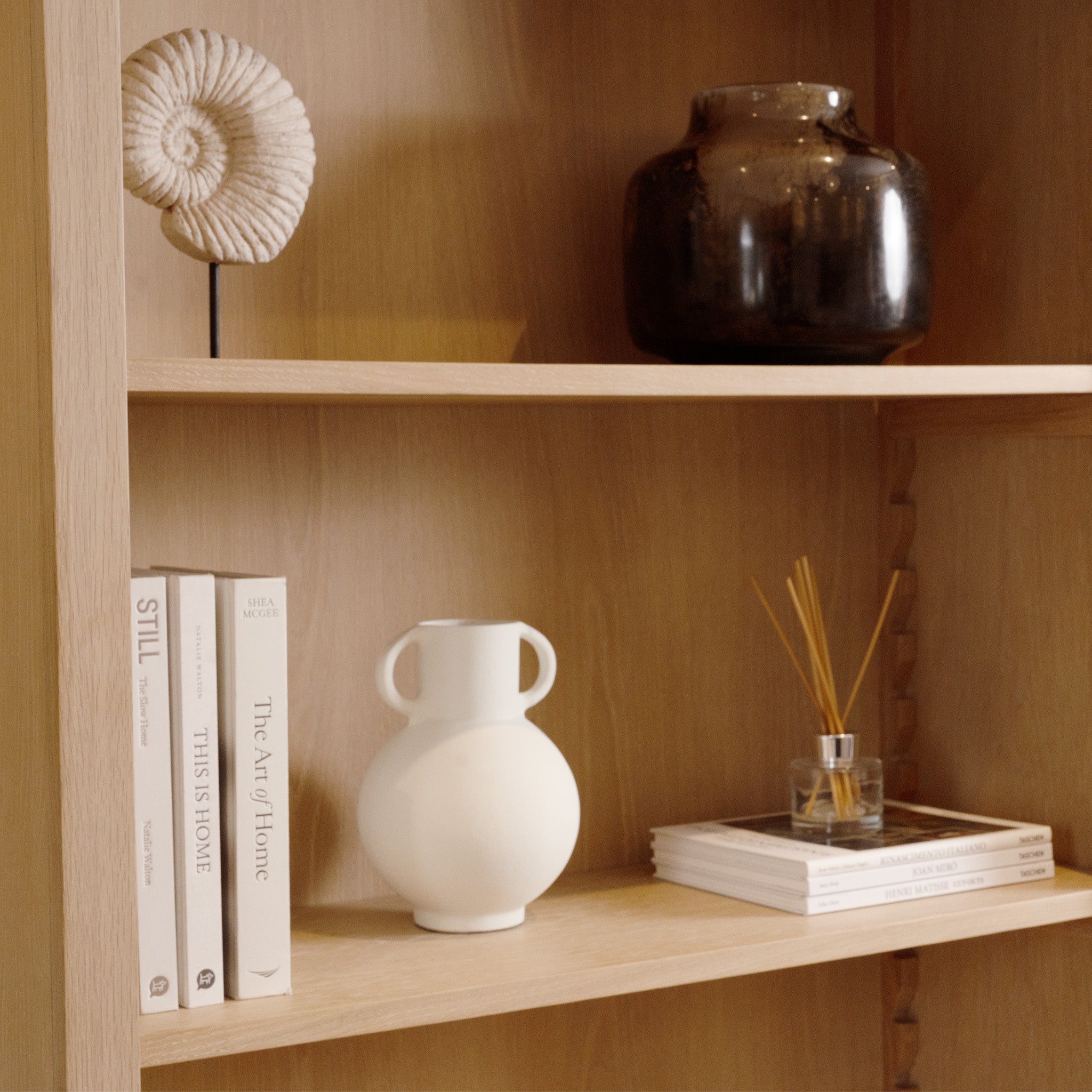 The Amberley Large Bookcase in Light Oak features a minimalist design, styled with books, a white vase, dark jar, seashell decor, and reed diffusers.