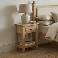 Amberley Bedside Table in Light Oak with a drawer, topped with a beige lamp and a bowl, placed beside a bed dressed in neutral bedding.