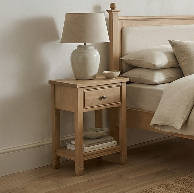 Amberley Bedside Table in Light Oak with a drawer, topped with a beige lamp and a bowl, placed beside a bed dressed in neutral bedding.
