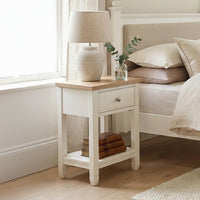 The Amberley Bedside Table in Warm White brings modern country charm to your bedroom, styled with a lamp, a small vase with leaves, and books stacked on the lower shelf beside the bed.