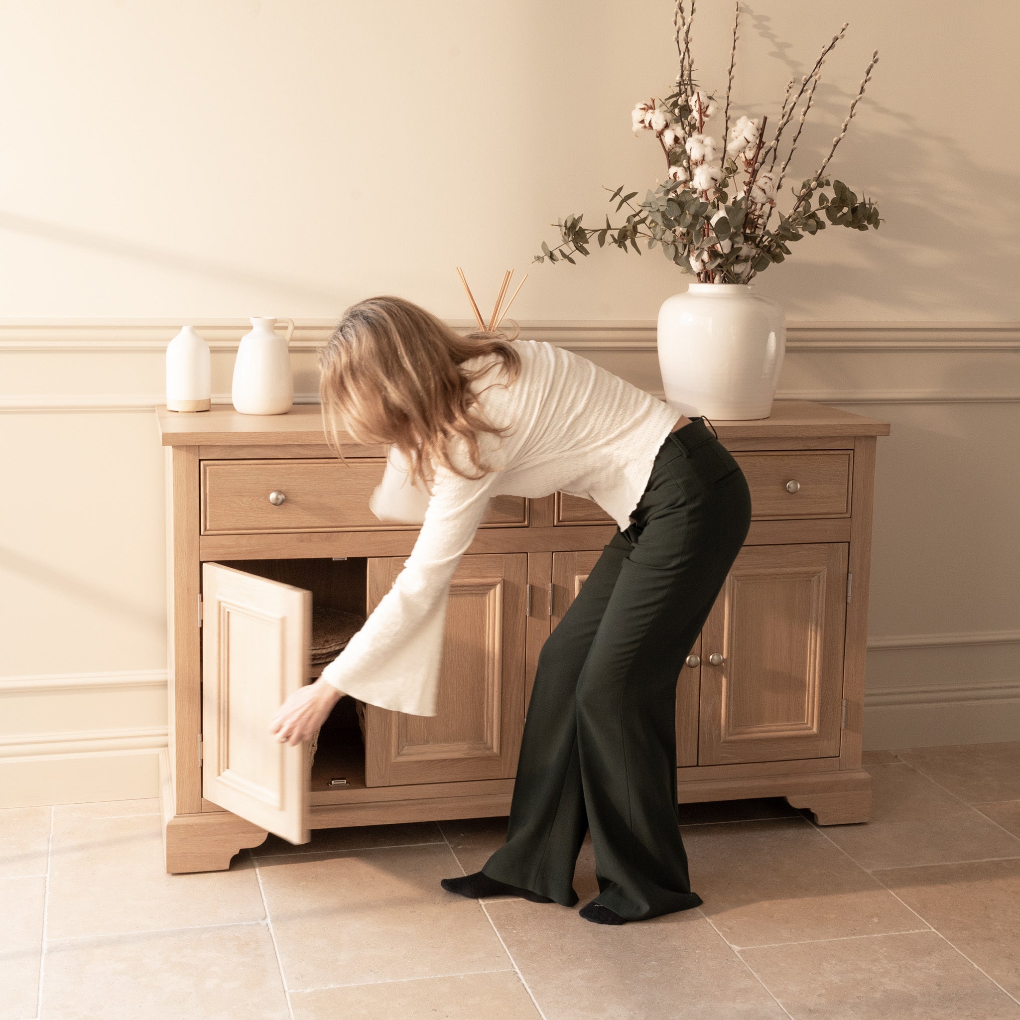 A woman opens the lower cabinet door of the Amberley Large 4 Door 2 Drawers Sideboard in Light Oak, which is decorated with vases and a plant.