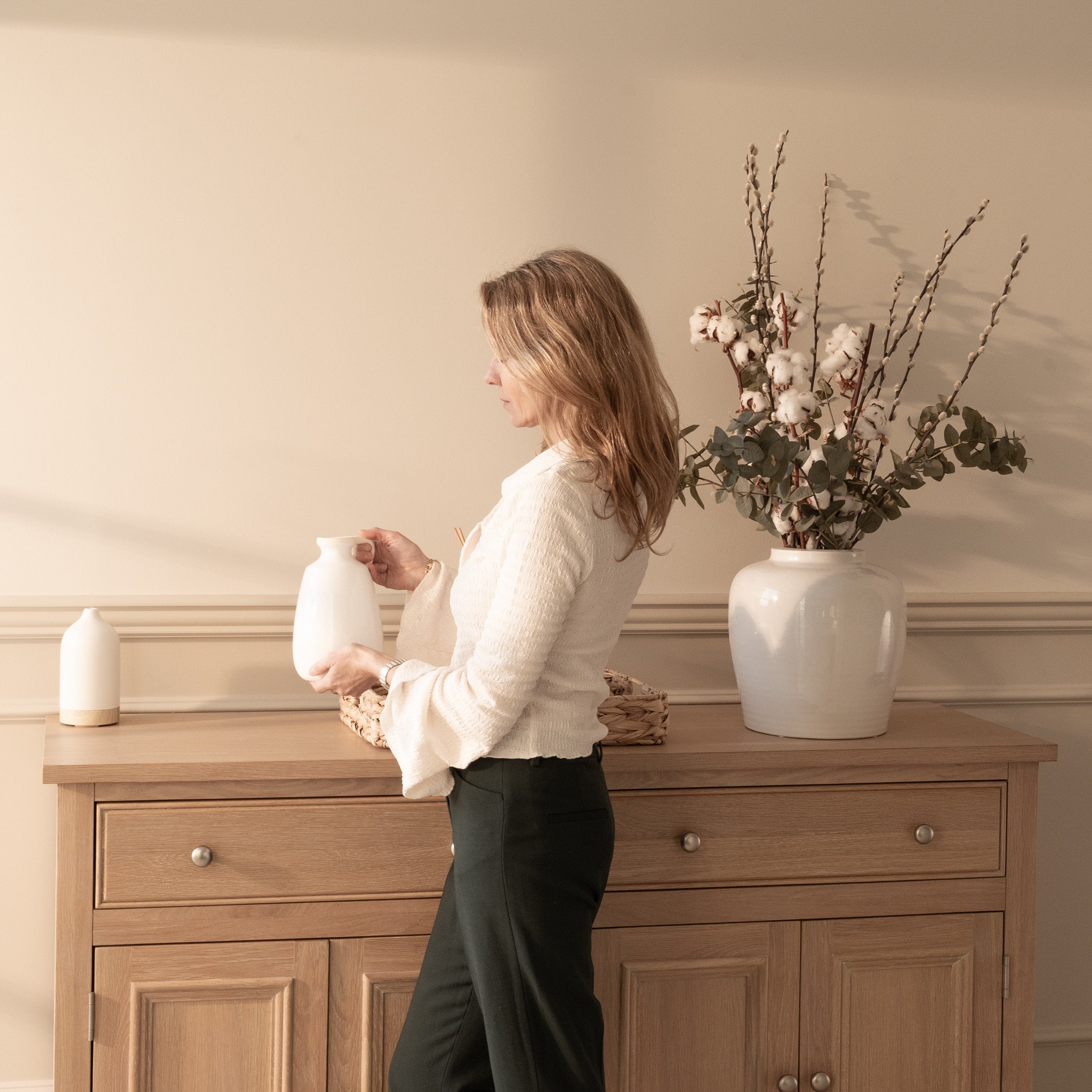 A woman holding a white vase stands beside the Amberley Large 4 Door 2 Drawers Sideboard in Light Oak, decorated with vases and a floral arrangement.