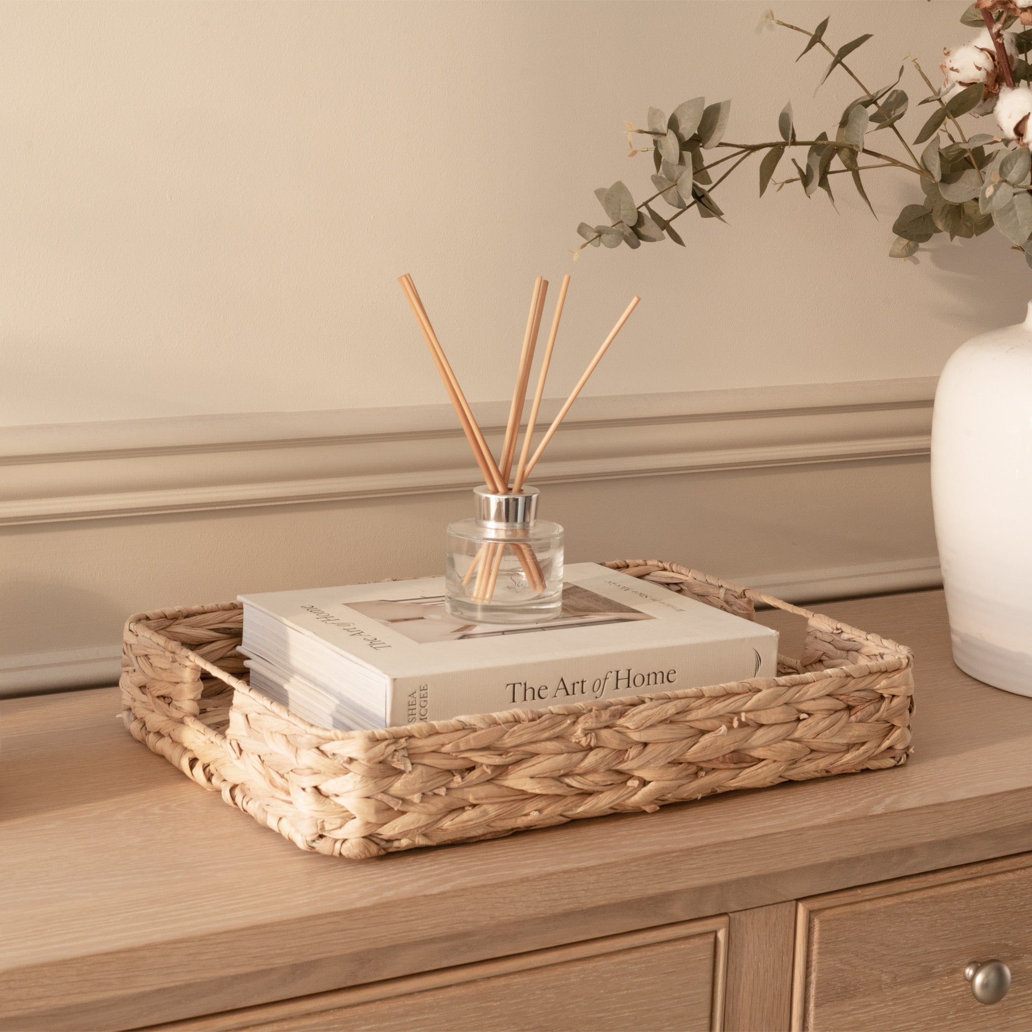 A woven tray with books and a reed diffuser sits on the Amberley Large 4 Door 2 Drawers Sideboard in Light Oak, next to a white vase with flowers.