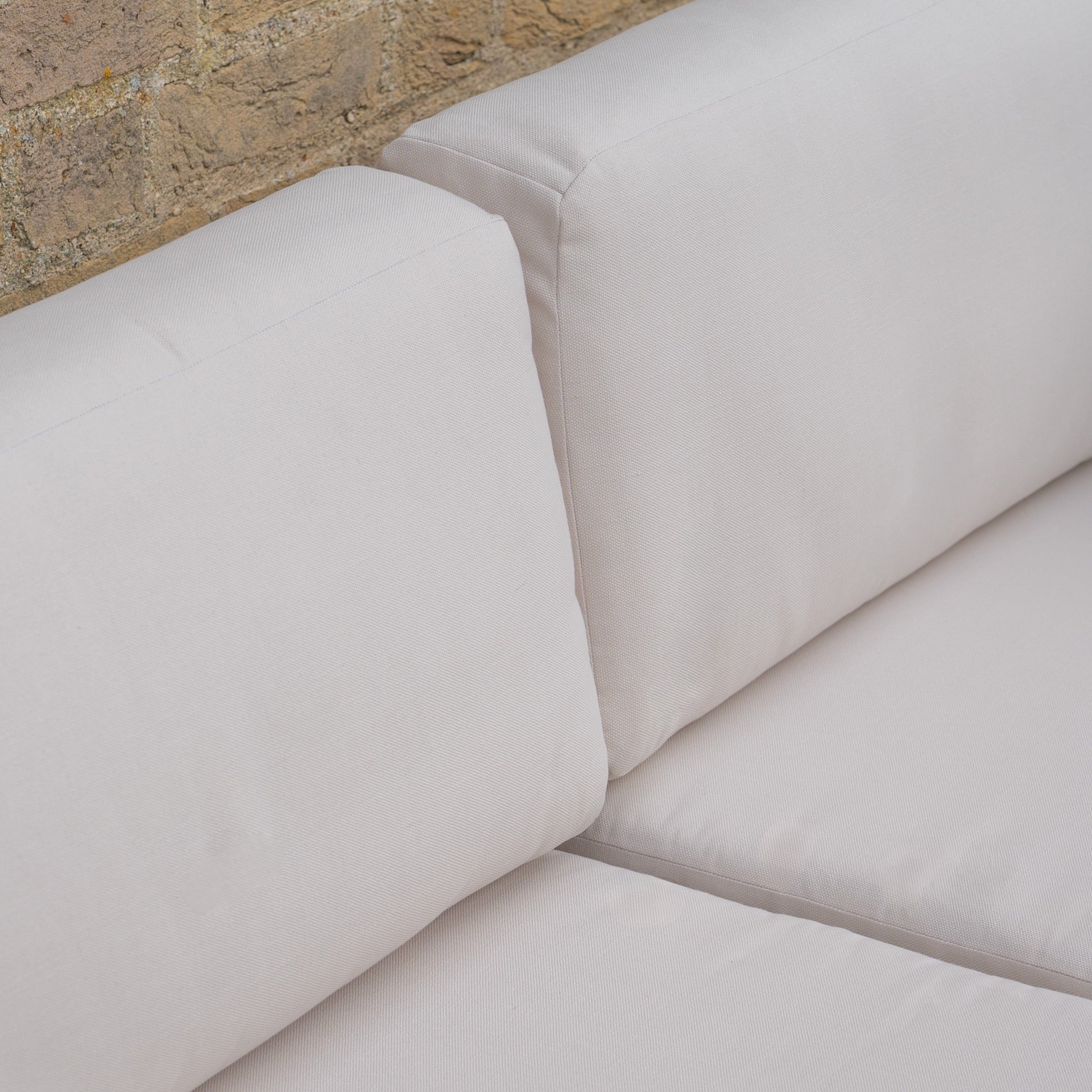 Close-up of the Andora Middle Piece in Weathered Brown and Linen, featuring light beige cushions against a brick wall—an outdoor modular piece crafted from weather-resistant eucalyptus wood.