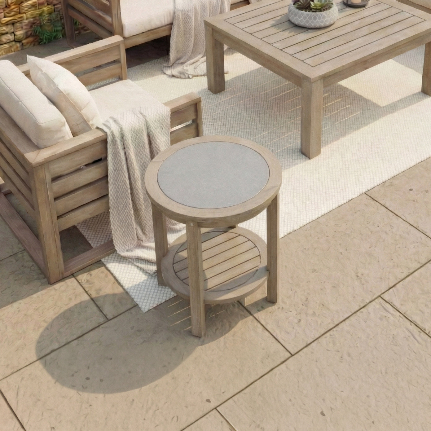 The Andora Round Side Table in Weathered Brown, crafted from eucalyptus wood, sits on a patio beside outdoor sofas and a round coffee table atop a white rug.