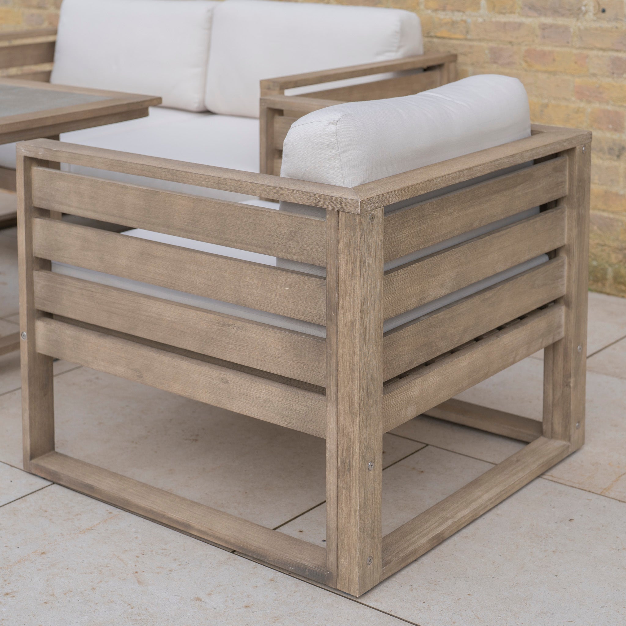 The Andora Armchair in Weathered Brown and Linen, featuring weather-resistant eucalyptus wood and light cushions, offers stylish garden seating on a patio beside a brick wall.
