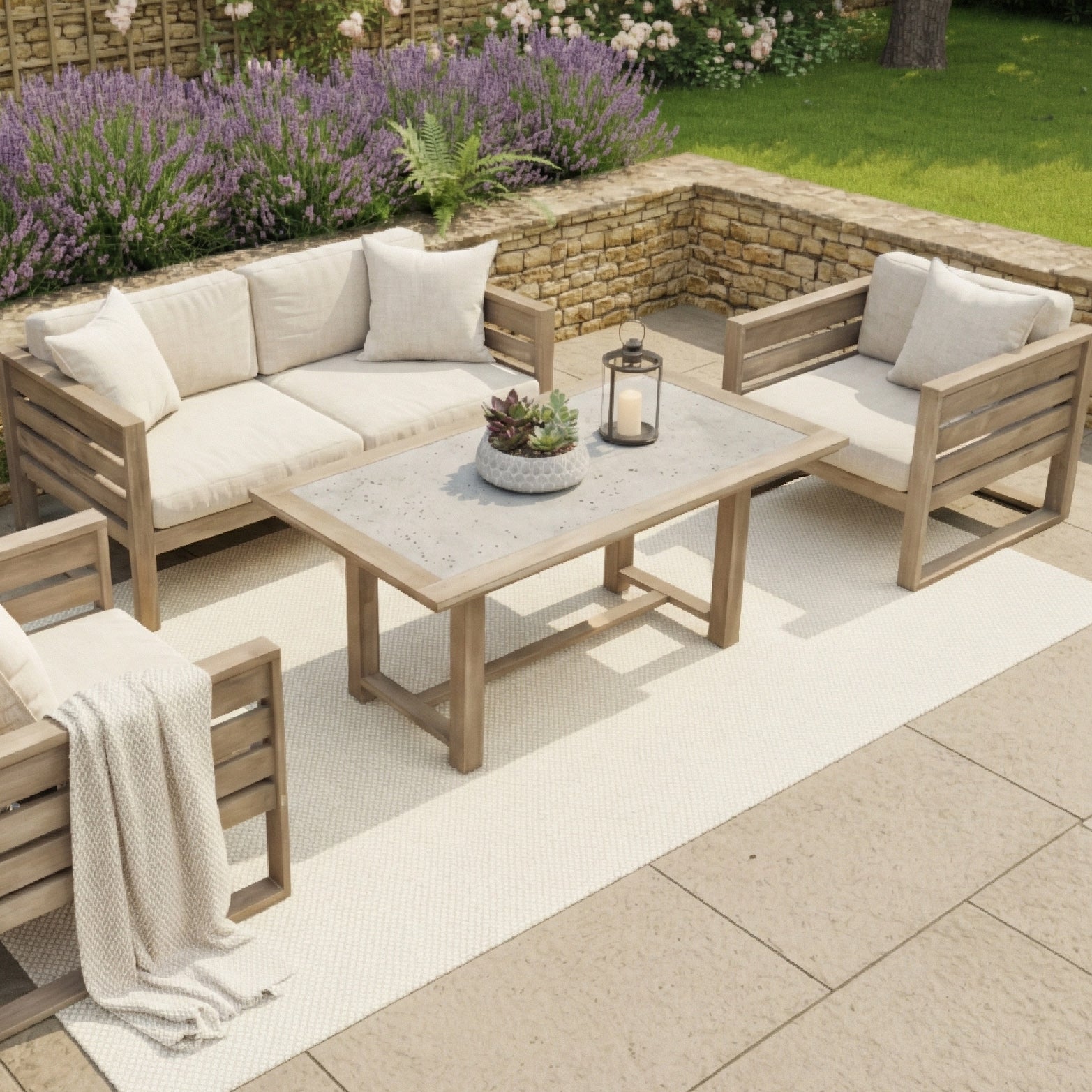 Wooden patio furniture set with beige cushions on a rug, featuring the Andora Coffee Table (mid-height, weathered brown), surrounded by stone walls and blooming garden plants.