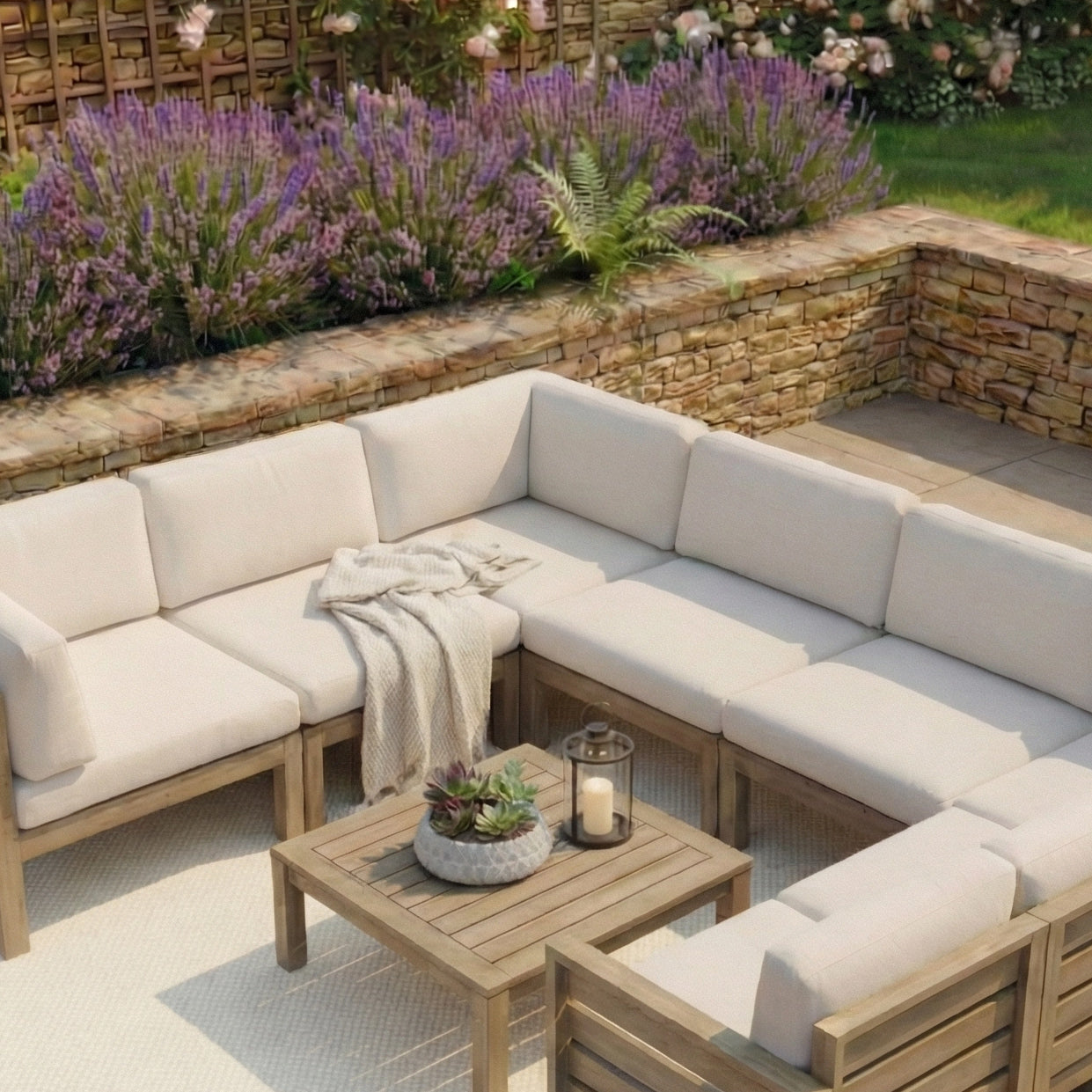 Outdoor patio featuring the Andora Middle Piece in Weathered Brown and Linen, paired with a throw blanket, wooden table, candle, and plants near a stone wall and lush garden.