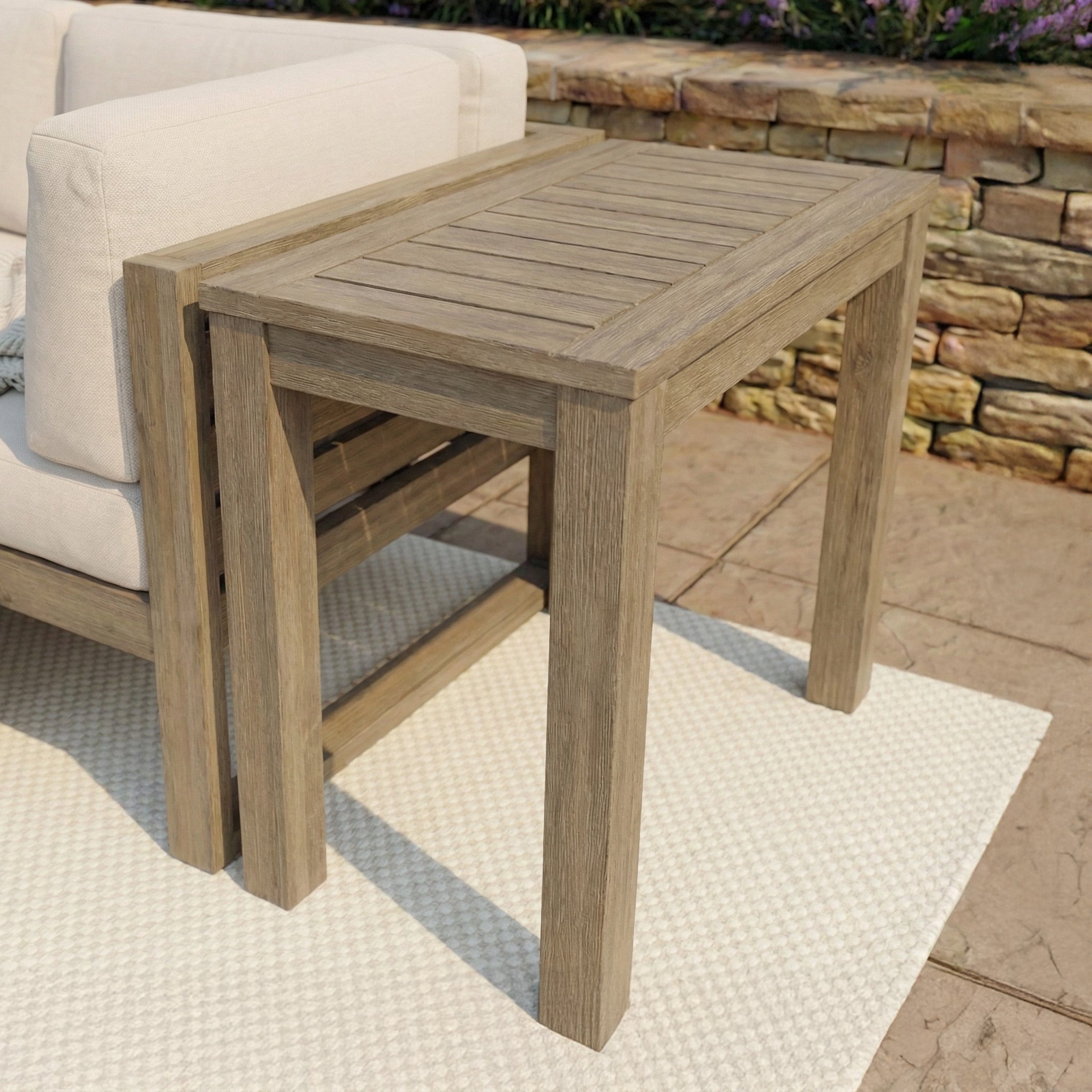 The Andora Narrow Side Table in Weathered Brown, crafted from solid eucalyptus wood, sits next to a patio sofa atop a light woven outdoor rug.