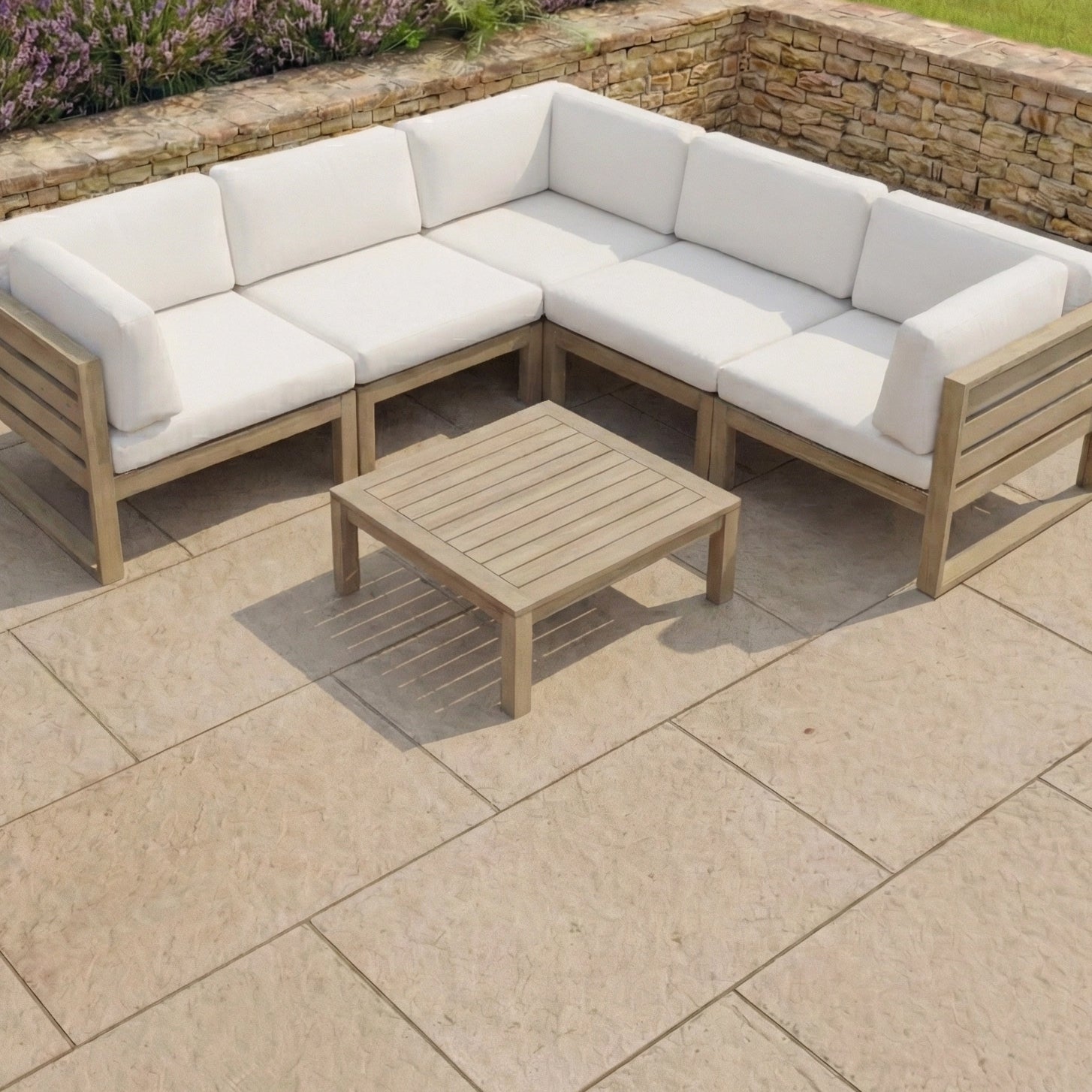Beige outdoor sectional sofa with white cushions and the Andora Coffee Table & Footstool in Weathered Brown and Linen, arranged on a stone patio near a stone wall.