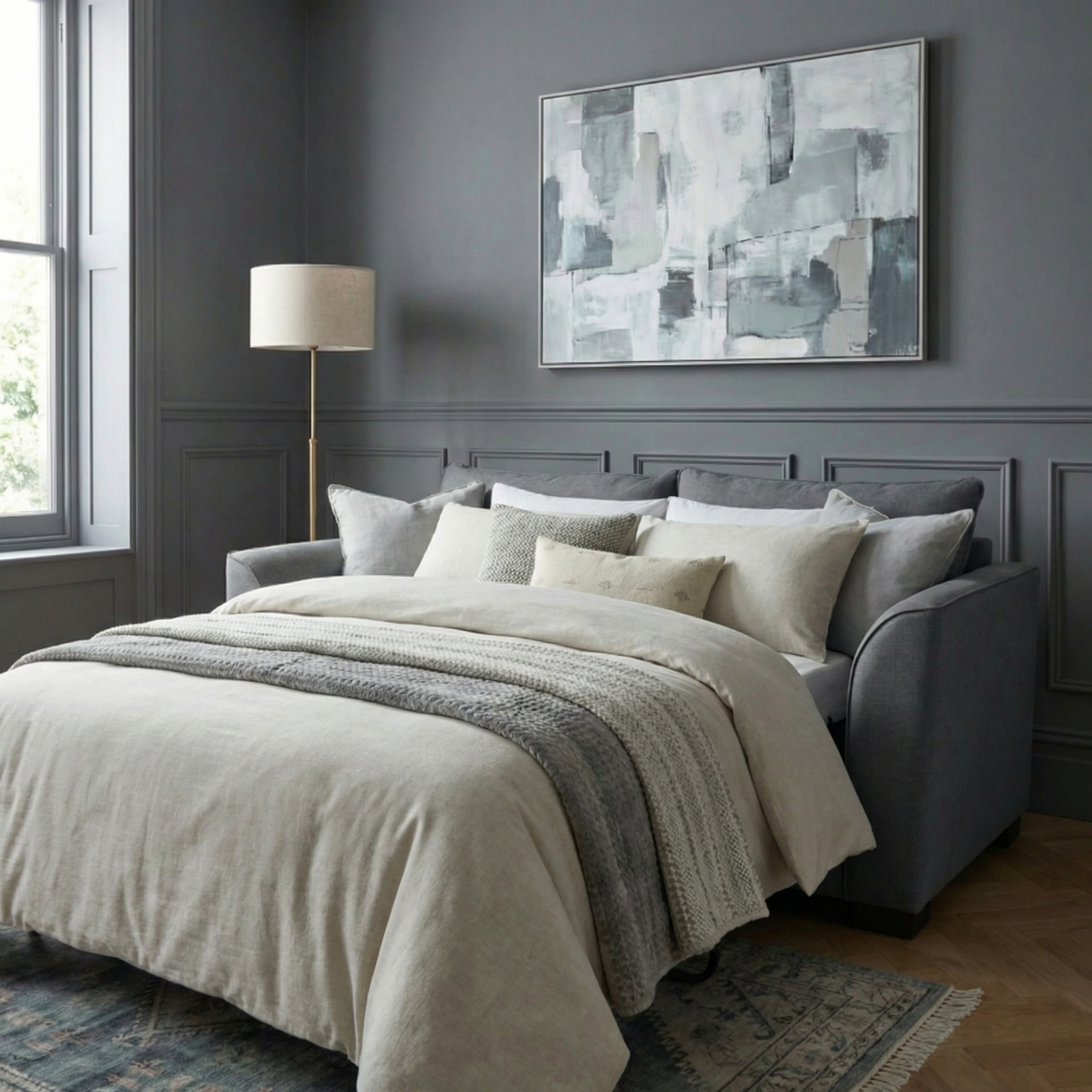 A modern bedroom featuring the Ardley 3 Seat Sofa Bed in Smoke Grey, complemented by neutral bedding, a standing lamp, and abstract wall art.