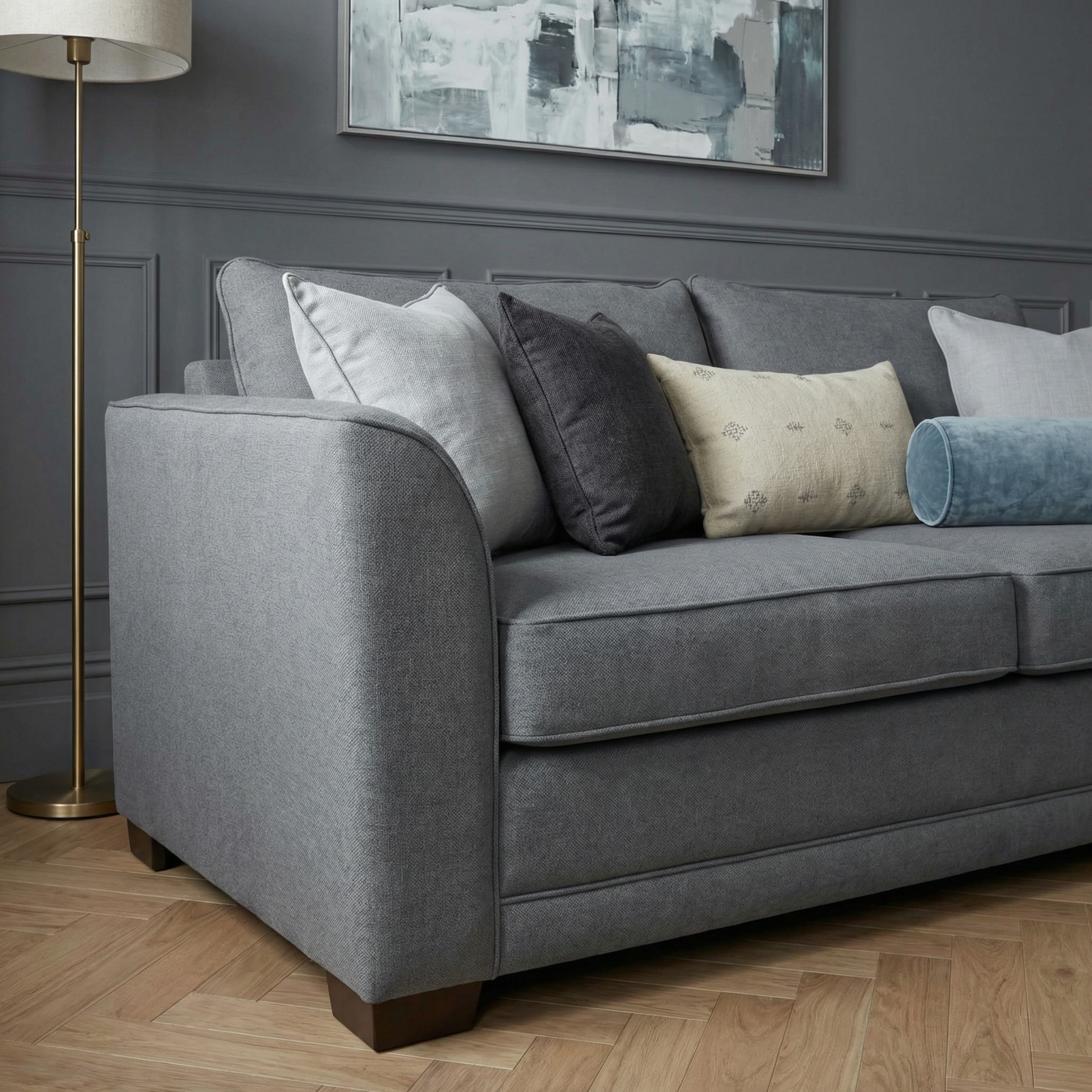 The Ardley 3 Seat Sofa Bed in Smoke Grey, made in Britain, features assorted pillows and a pocket-sprung mattress, perfect for modern living rooms with wood floors and abstract wall art.