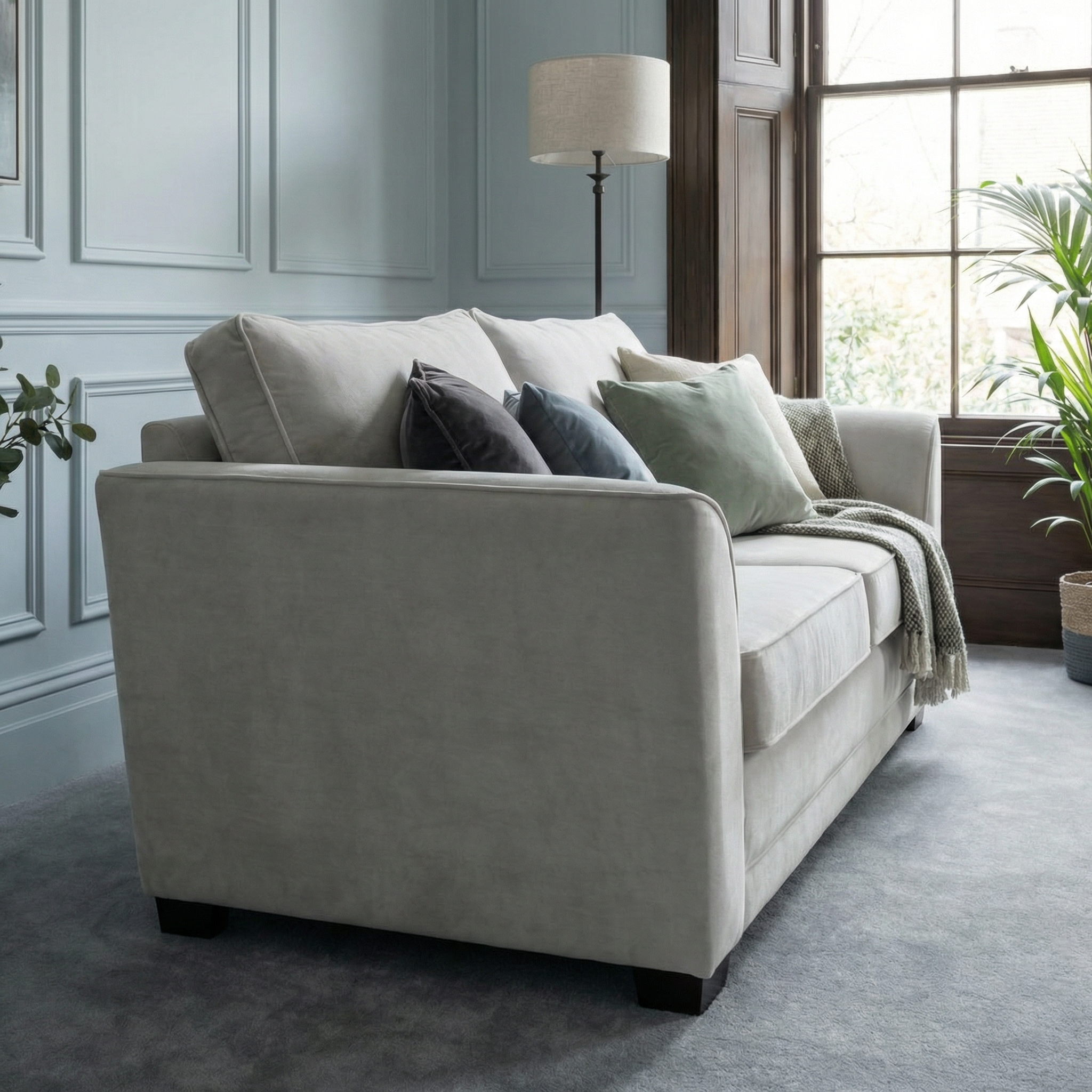 The Ardley 3 Seat Velvet Sofa Bed in Feather Grey, adorned with decorative pillows and a throw blanket, is placed in a room with a large window.
