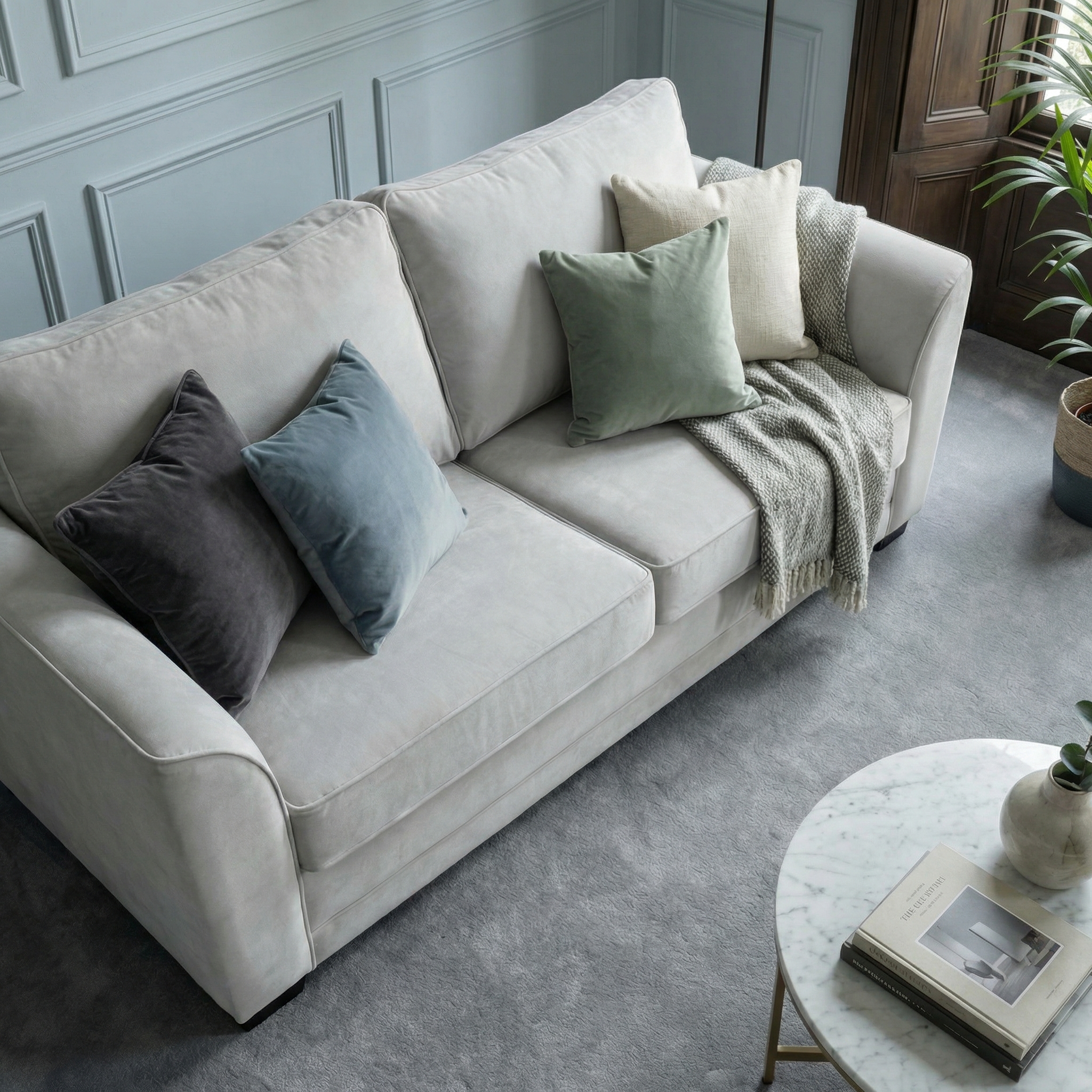 The Ardley 3 Seat Velvet Sofa Bed in Feather Grey, adorned with mixed throw pillows and a blanket, sits beside a round marble coffee table holding a book and plant.