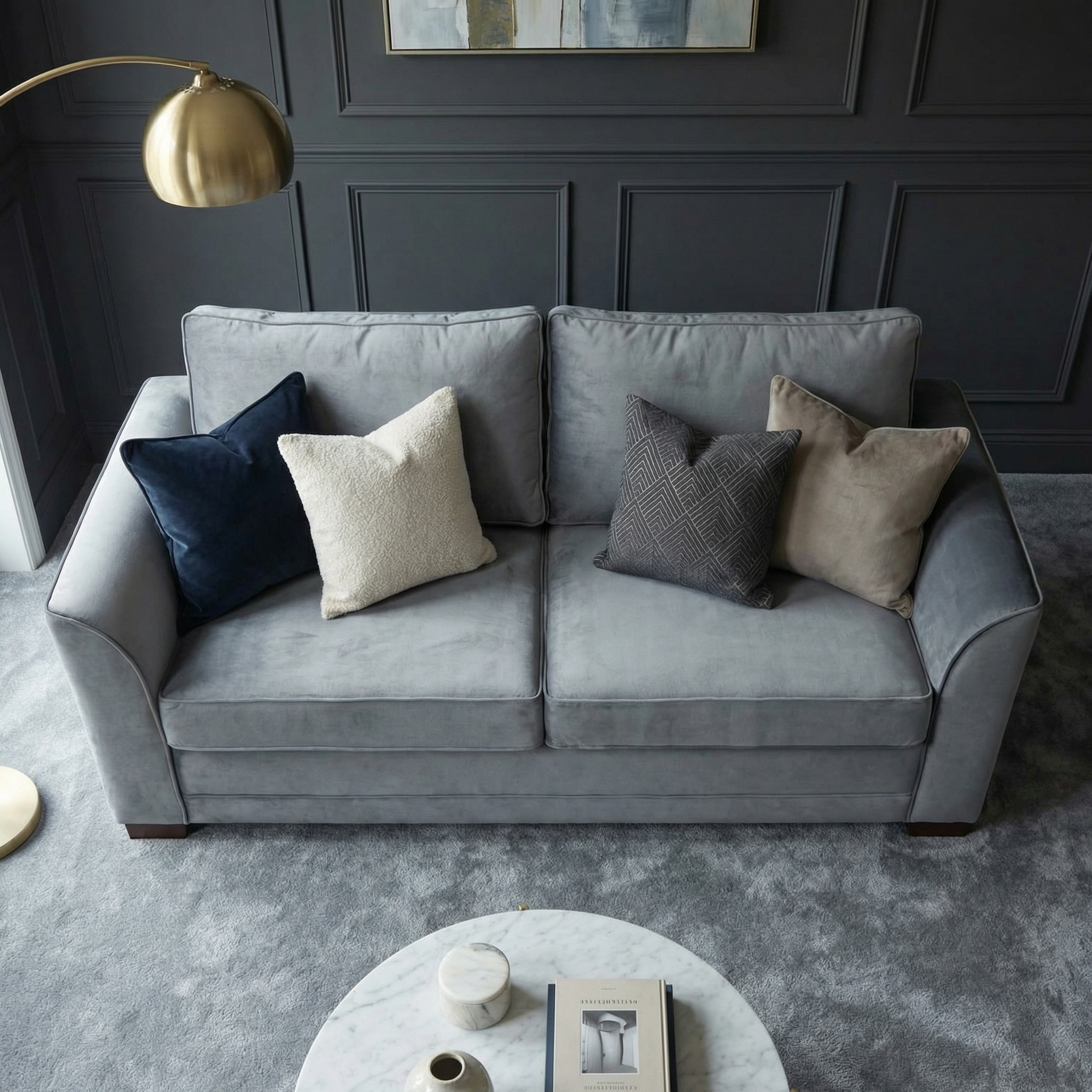 The Ardley 3 Seat Velvet Sofa Bed in Steel Grey is shown with four assorted throw pillows, placed next to a gold floor lamp on a gray carpeted floor.
