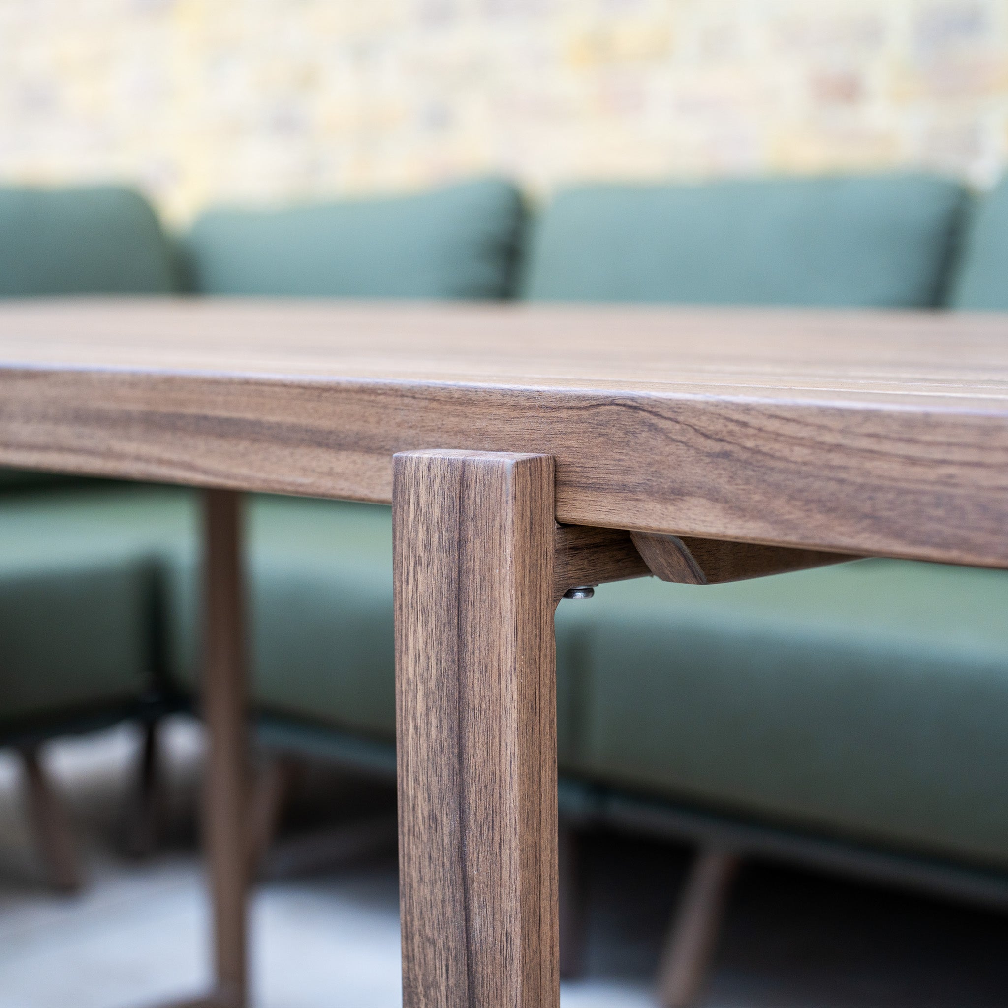 Close-up of a wooden table edge from the Ascot Corner Dining Set with Armchair in Forest Green, featuring green cushions and a brick wall—perfect for stylish garden furniture or any outdoor dining arrangement.