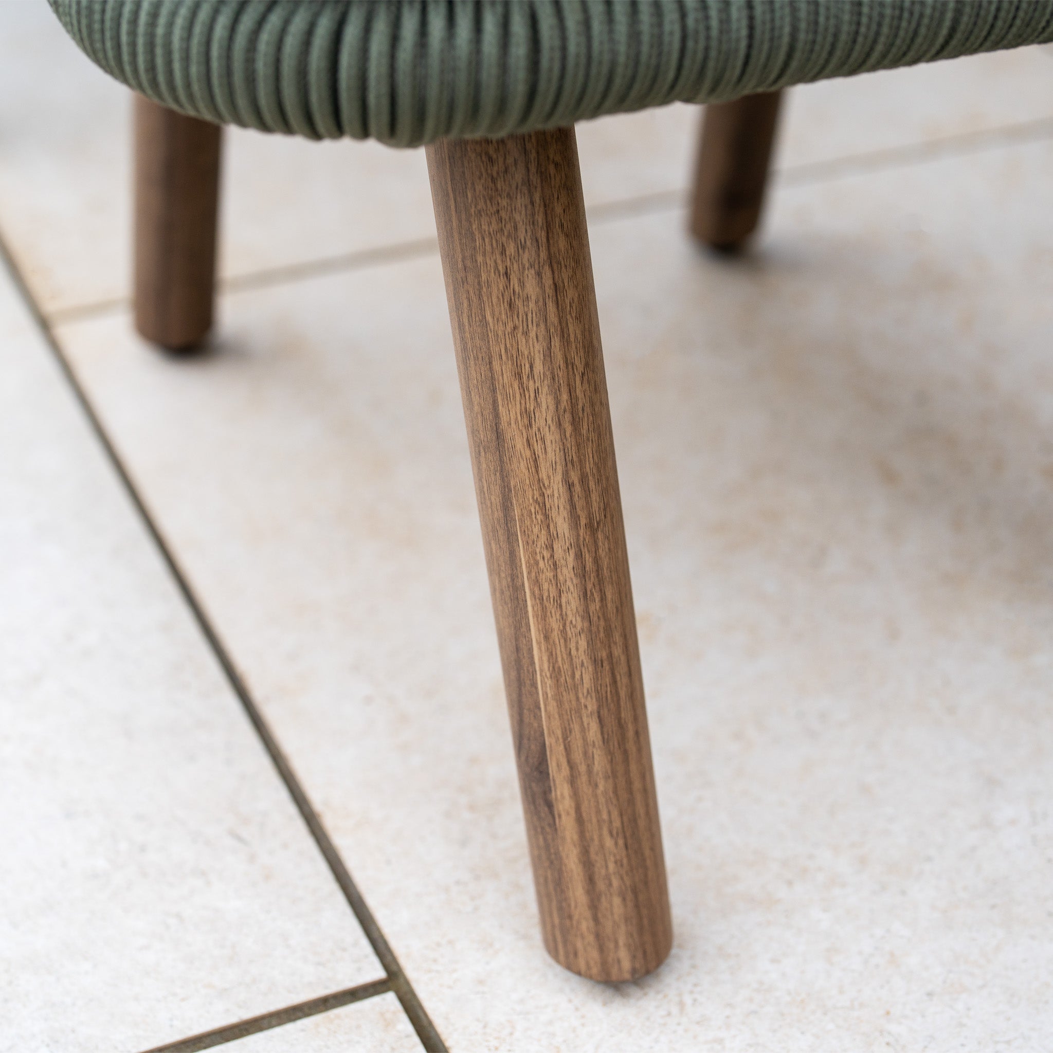 Close-up of a wooden chair leg on tiles with a hint of green upholstery—an inviting touch, echoing the weather resistance and comfort of the Ascot Corner Dining Set in Forest Green.