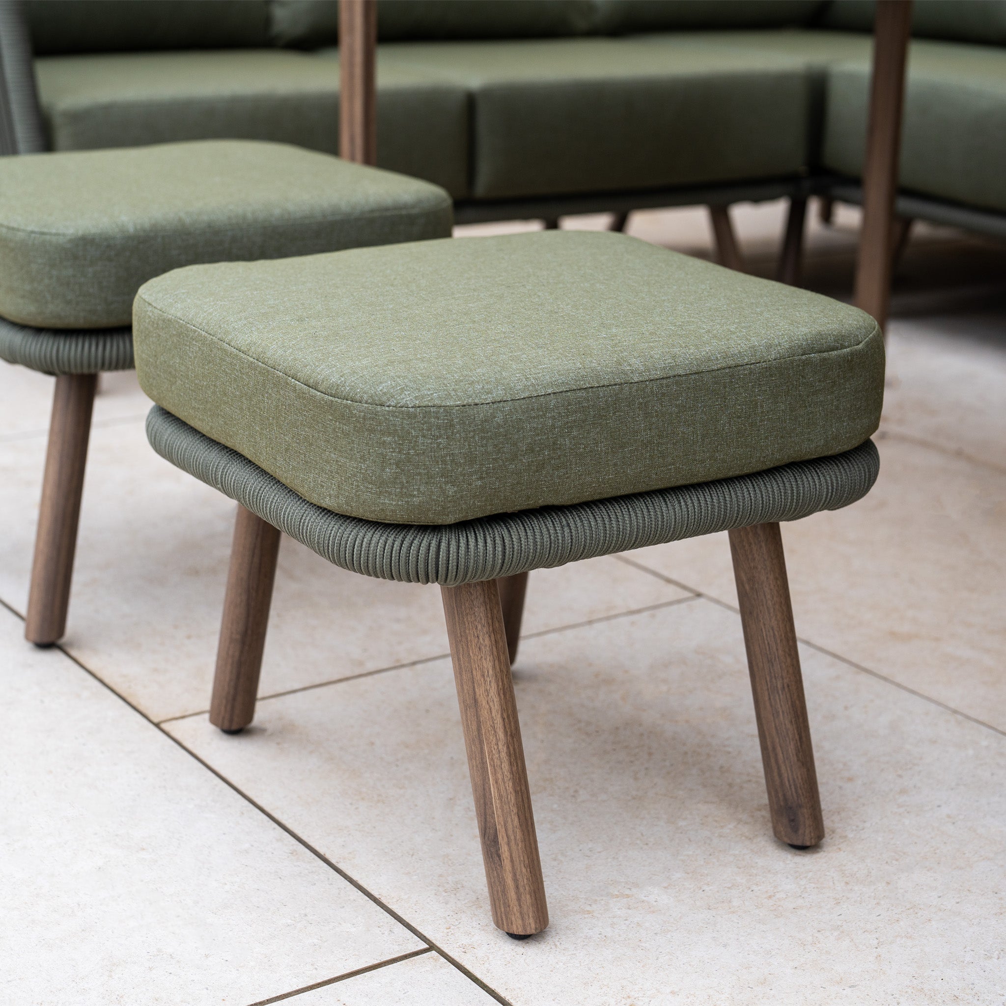 A green cushioned stool with wooden legs sits on a tiled floor, matching the Ascot Corner Dining Set with Armchair in Forest Green; another stool and a sofa appear in the background.