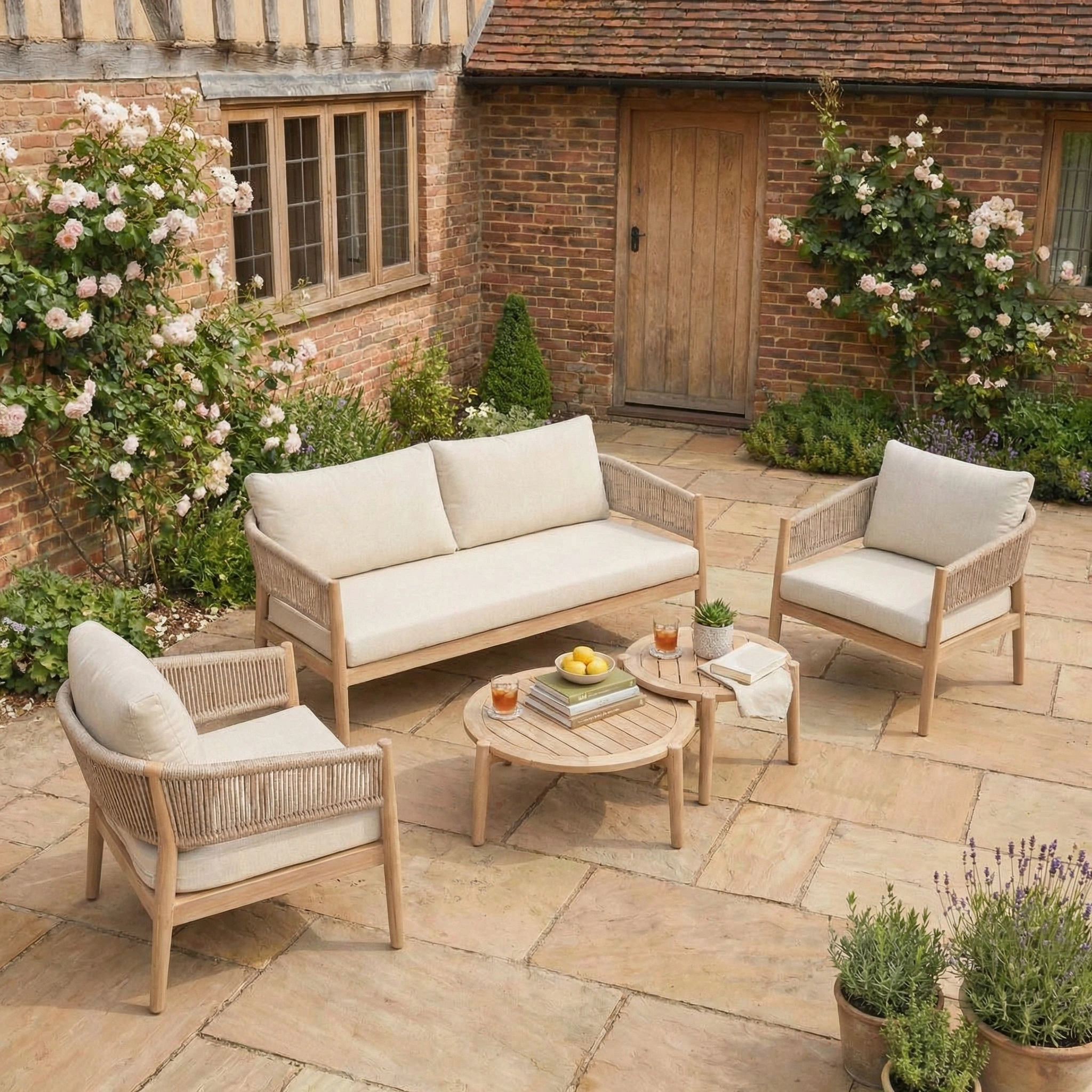 The outdoor patio features the Astoria 2 Seat Sofa Set in Ash Grey, with two chairs, a table set with drinks, and blooming plants beside a brick house.