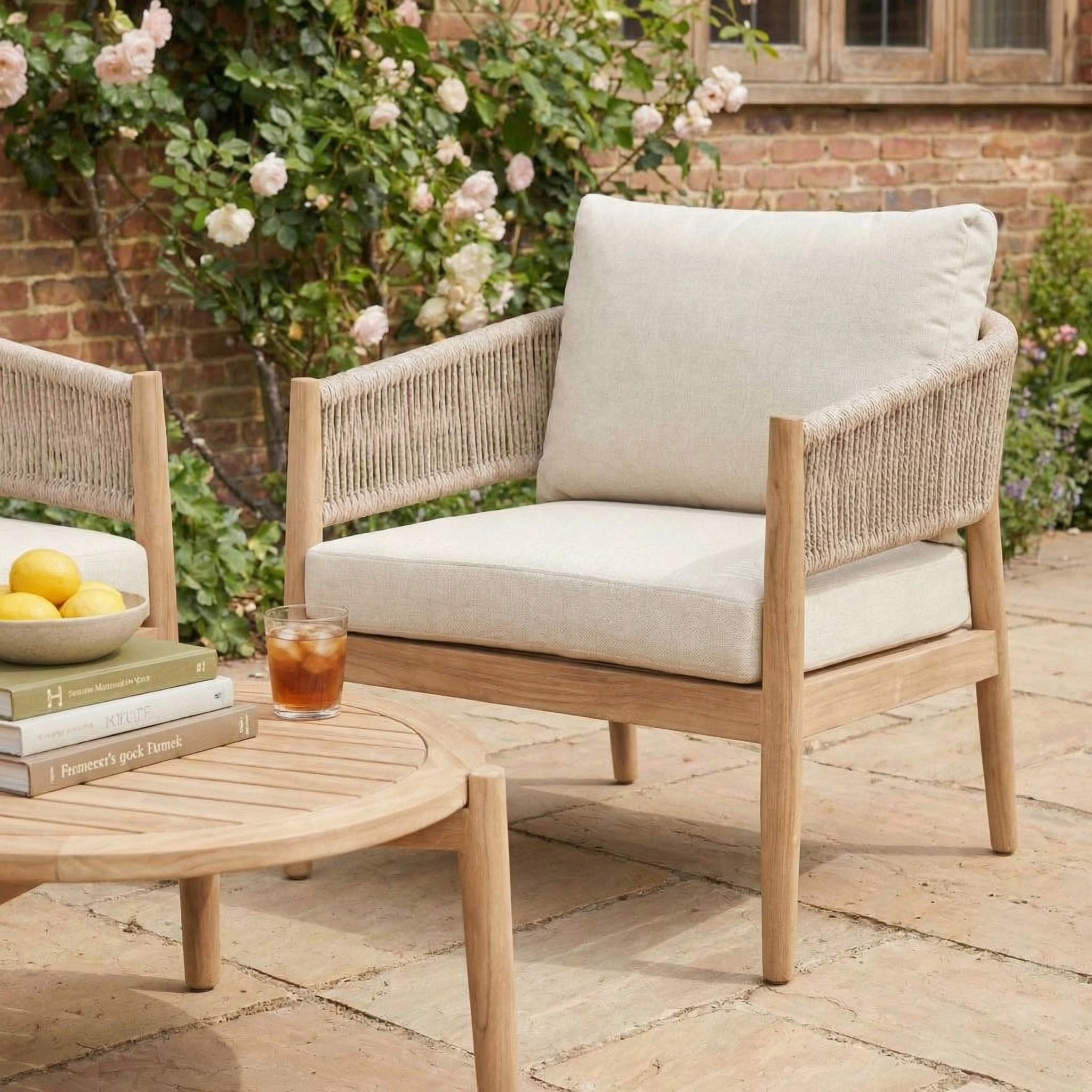 The Astoria 2 Seat Sofa Set in Ash Grey pairs beautifully with wooden patio chairs and light cushions beside a round table with books, lemons, and a drink on a sunny terrace.
