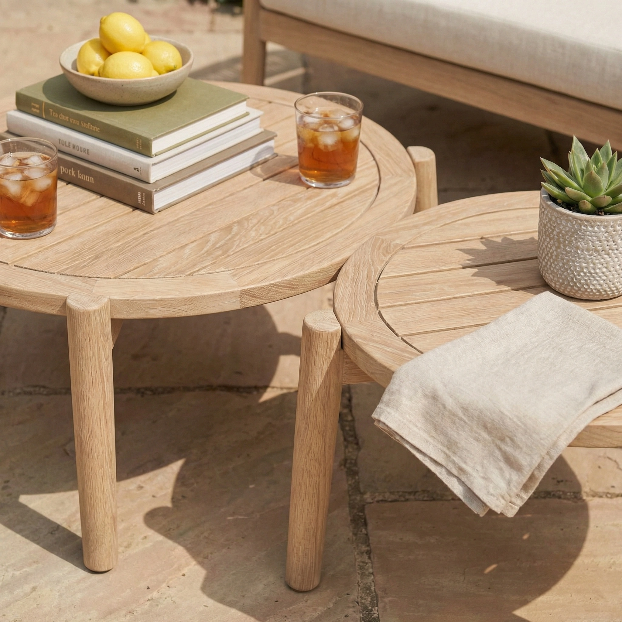 Two wooden tables with drinks, books, lemons, and a potted succulent sit beside the Astoria 2 Seat Sofa Set in Ash Grey on a stone patio.