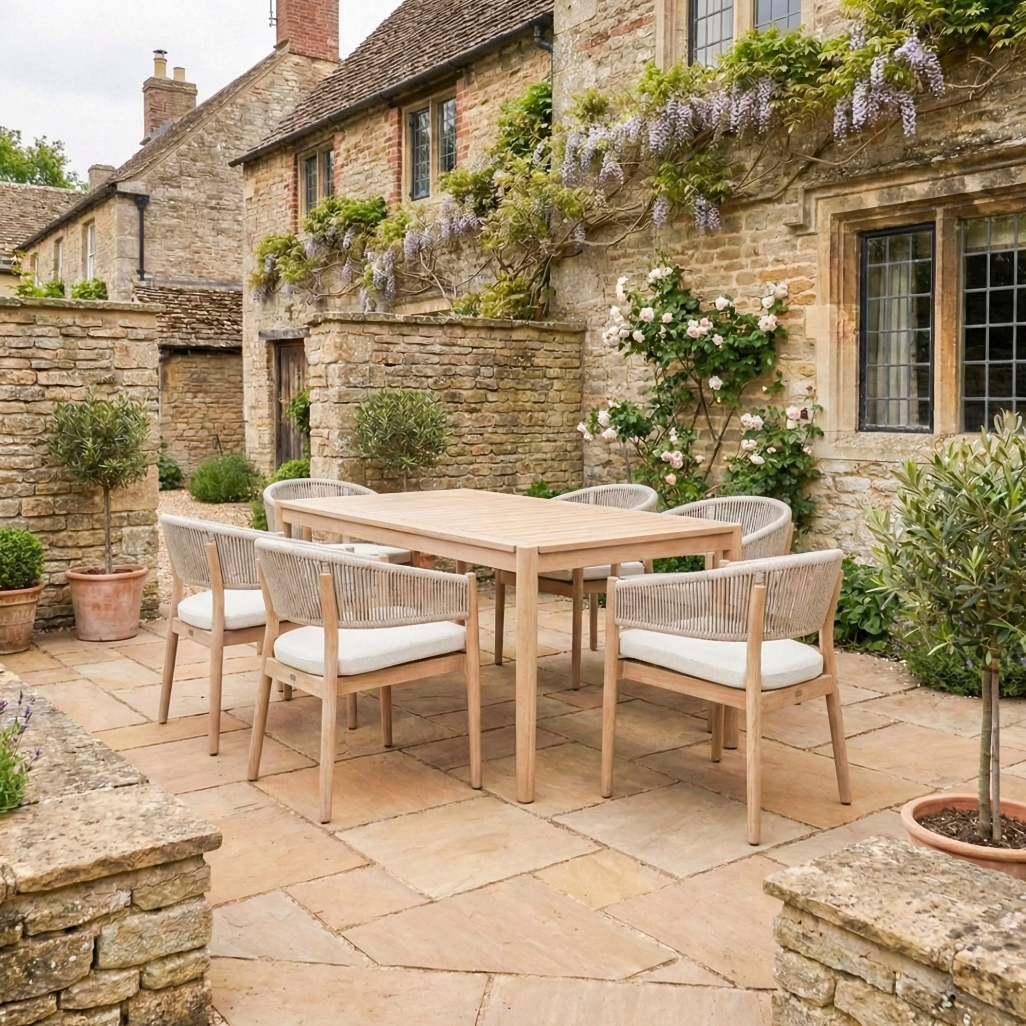 An Astoria 6 Seat Rectangular Dining Set in Ash Grey is placed on a rustic stone courtyard beside a charming house with flowers, seamlessly blending contemporary outdoor dining style with classic elegance.