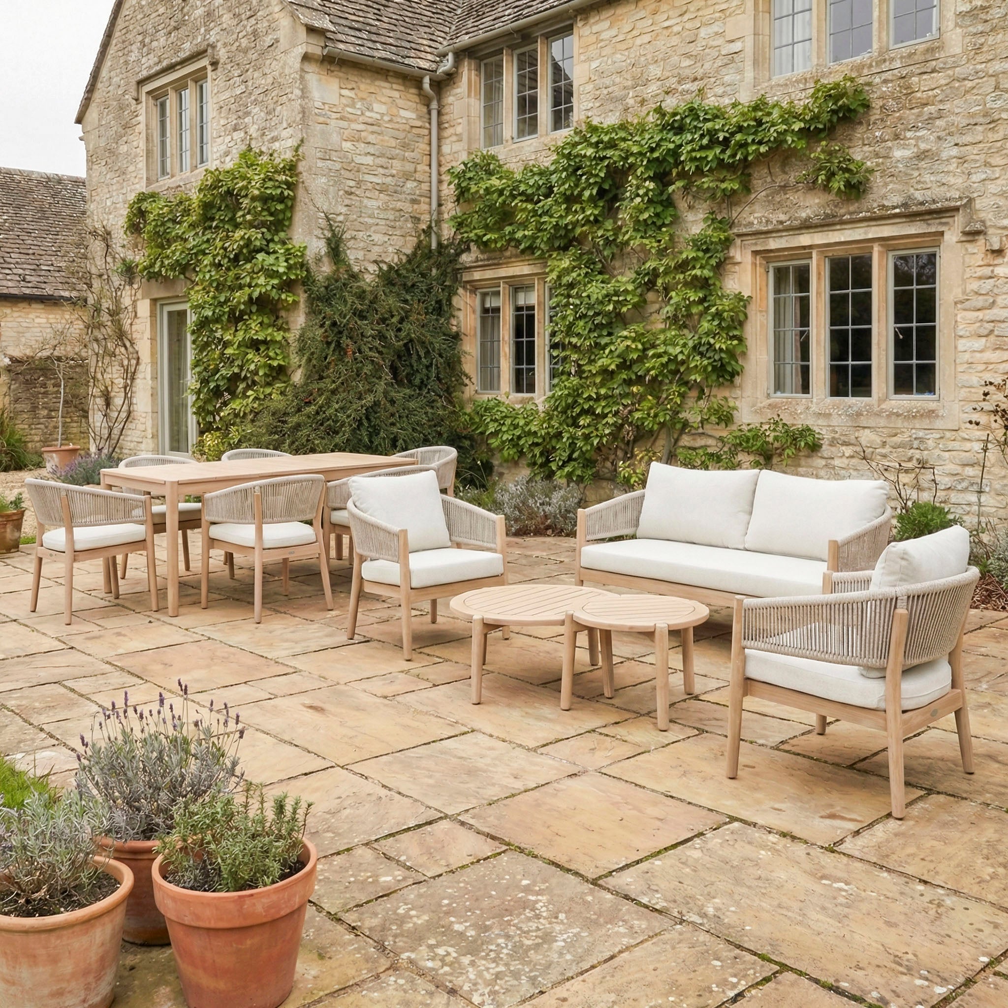 Outdoor patio with beige stone tiles, wooden furniture, potted plants, a stone house with green vines, and the Astoria 6 Seat Rectangular Dining Set in Ash Grey.