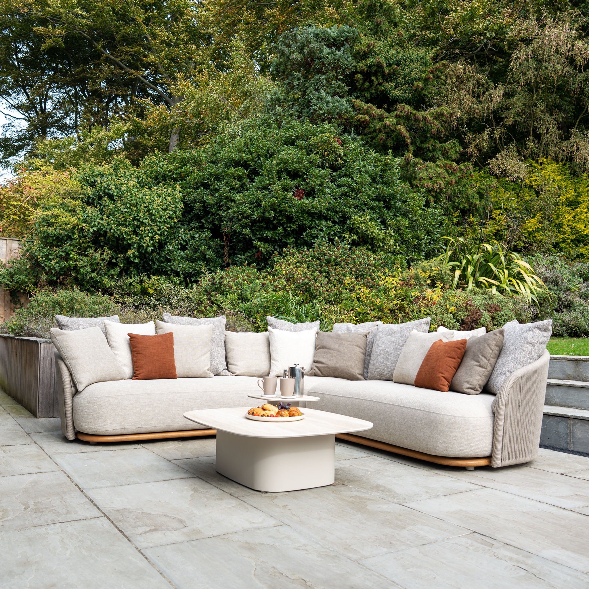 Modern outdoor seating area featuring the weather-resistant Avola Corner Sofa Set in Fawn, a stylish coffee table, and a lush green garden backdrop.