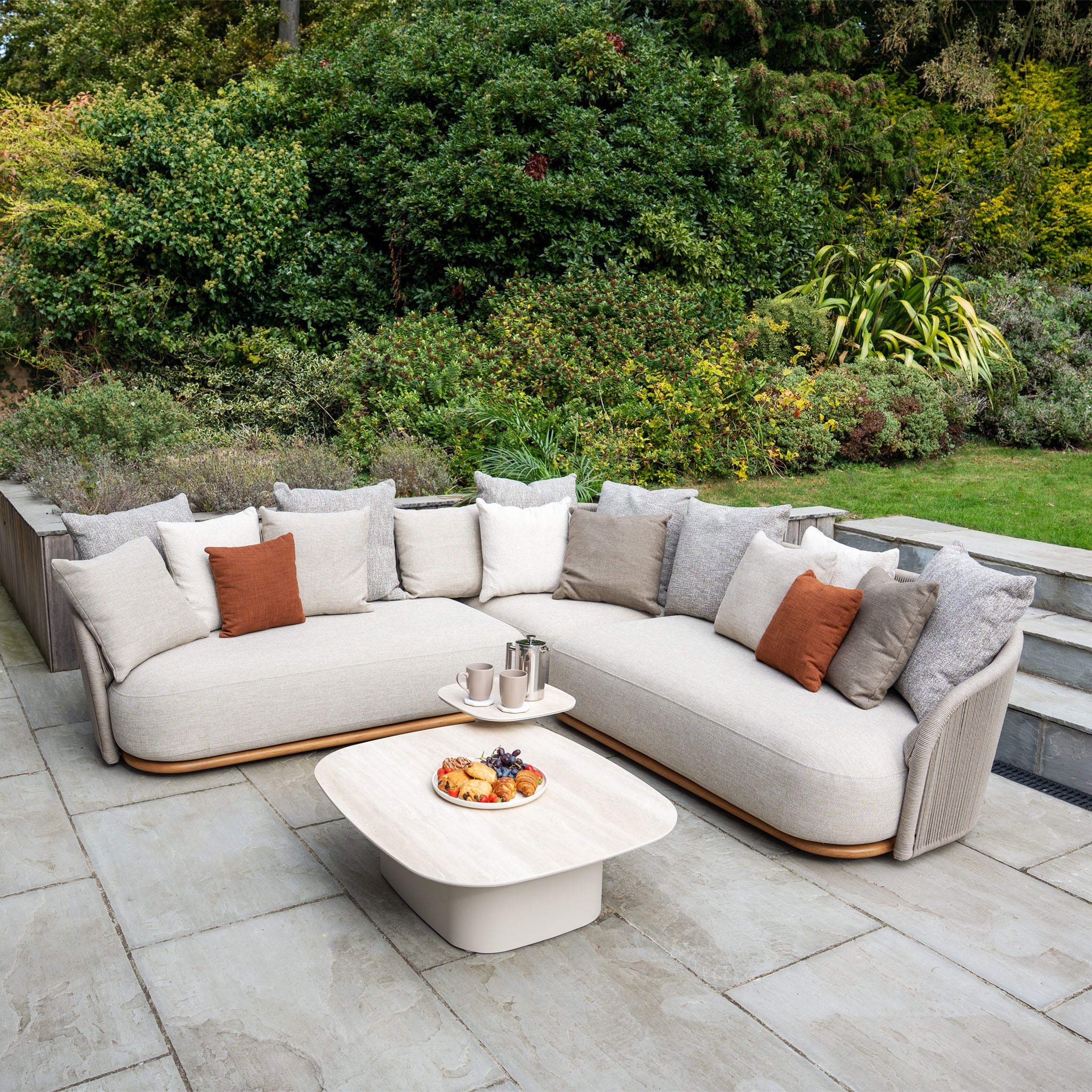 The Avola Corner Sofa Set in Fawn features a curved, weather-resistant outdoor sofa with cushions and a coffee table, perfect for relaxing on a patio surrounded by greenery.