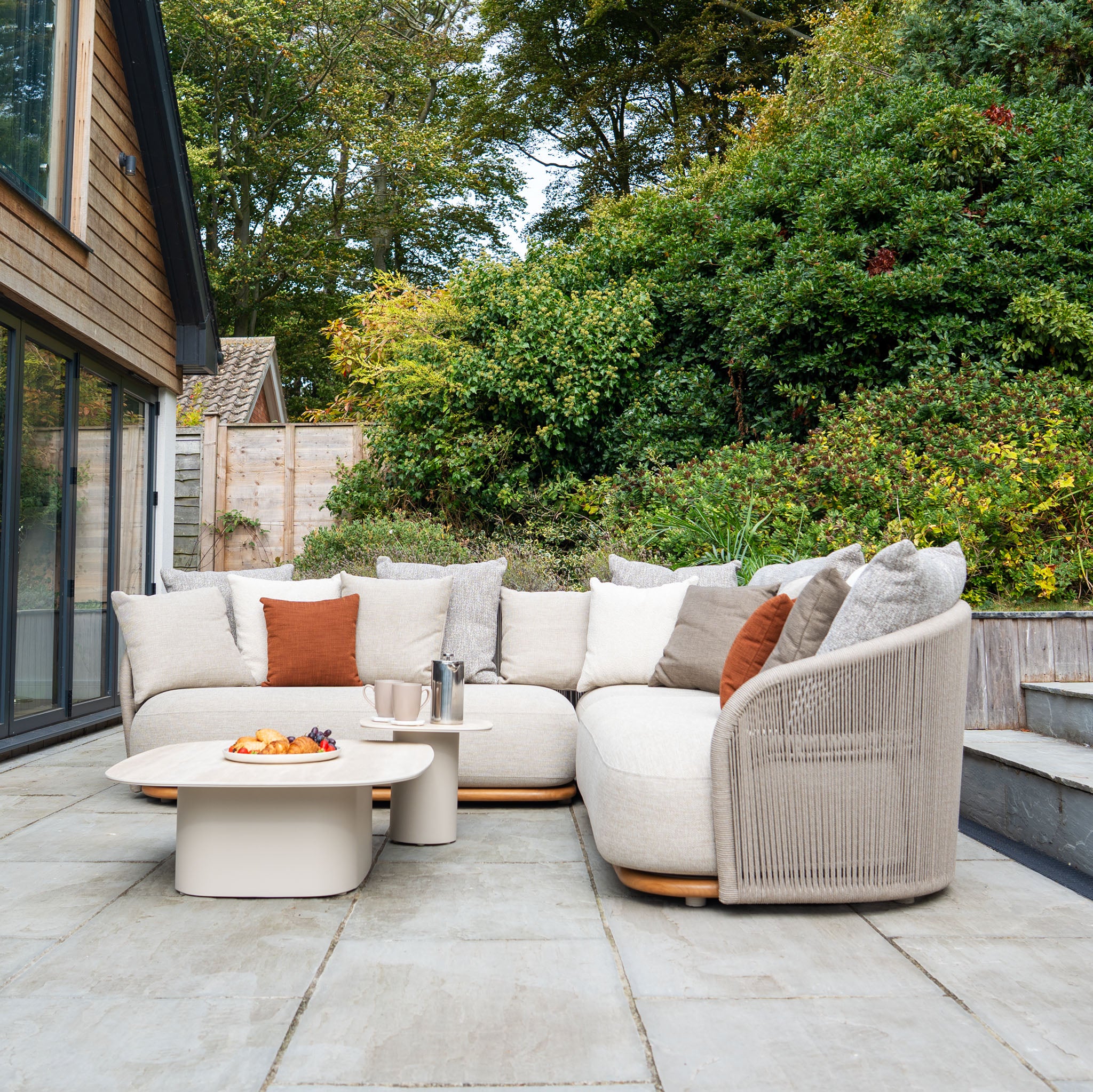 Outdoor patio with a weather-resistant Avola Corner Sofa Set in Fawn, coffee tables, and snacks, nestled among greenery beside a wooden house.