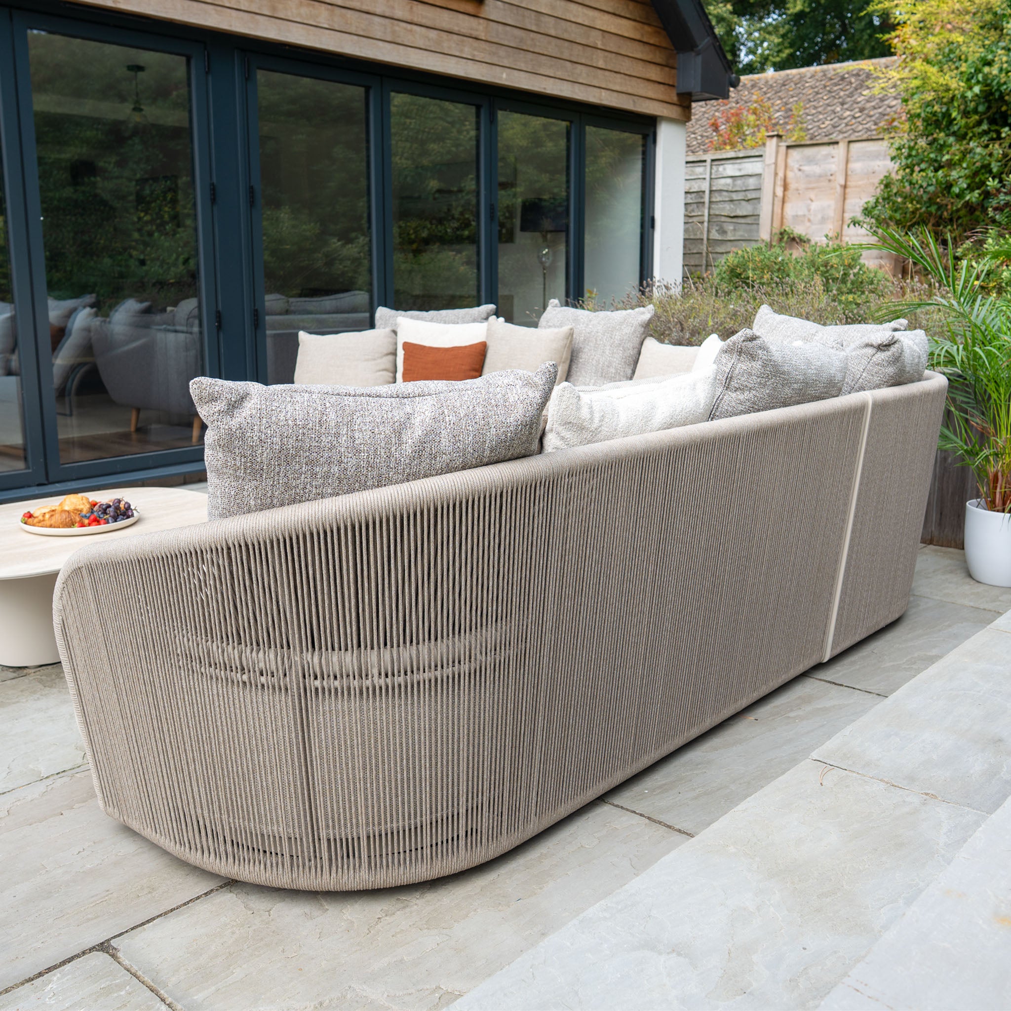 The Avola Corner Sofa Set in Fawn features neutral, weather-resistant olefin cushions and is perfect for stone patios by glass doors and potted plants, creating an ideal outdoor garden sofa scene.