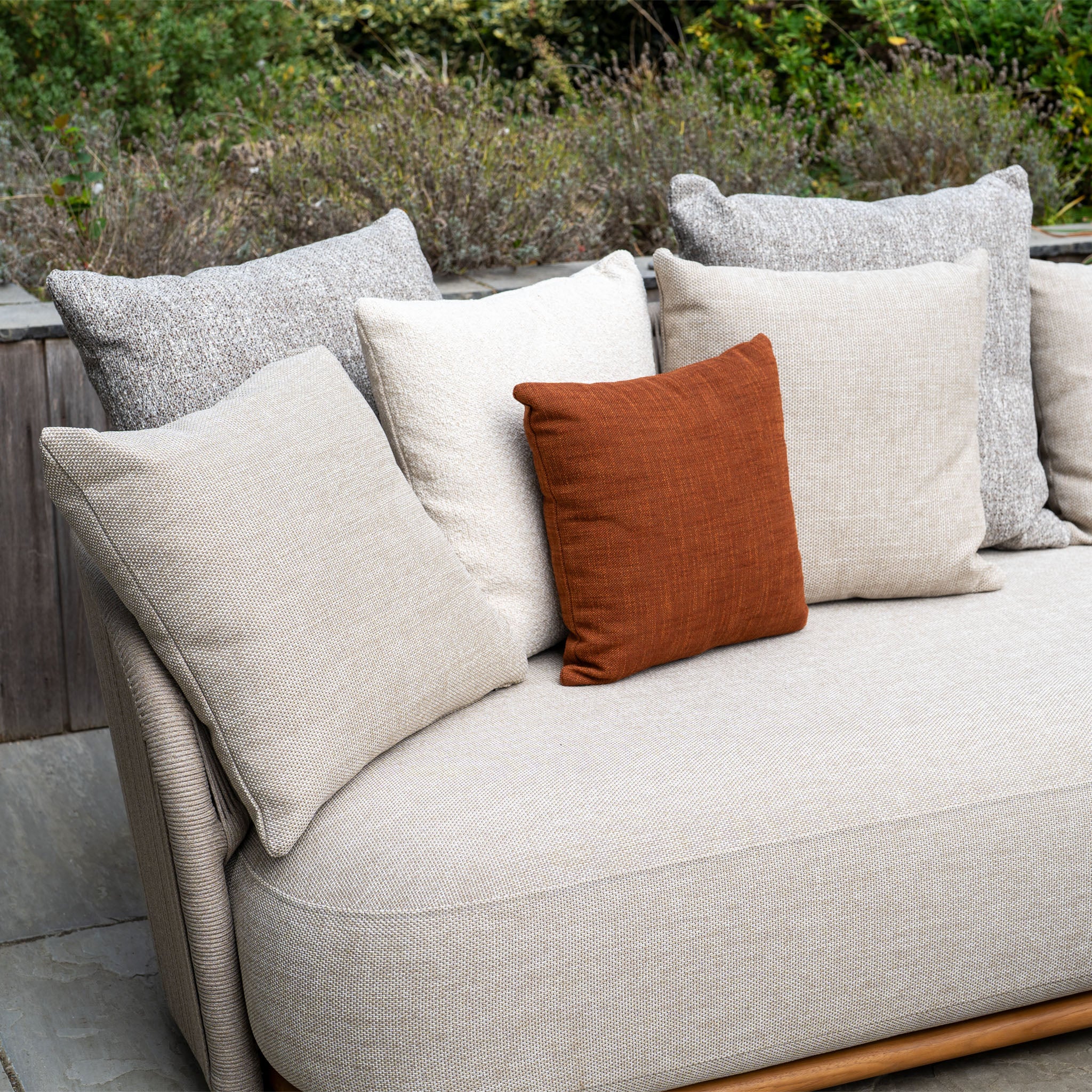 The Avola Corner Sofa Set in Fawn features weather-resistant olefin and six cushions, including a standout rust-colored one, perfect for relaxing outdoors amid greenery.