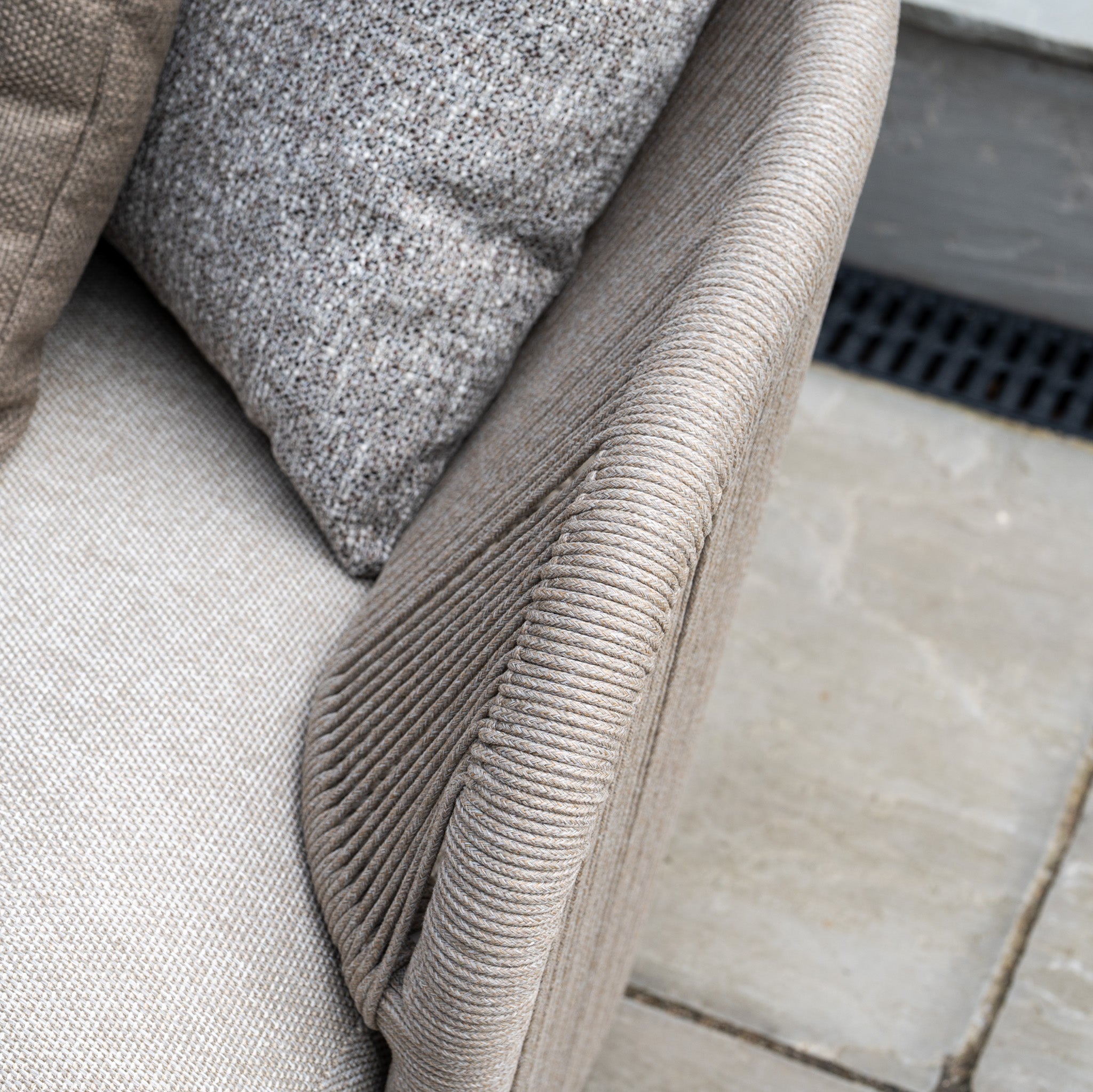 Close-up of a beige textured armchair with a gray cushion, part of the Avola Corner Sofa Set in Fawn, on a stone tile floor. Crafted from weather-resistant olefin, this outdoor garden sofa offers lasting style and comfort.