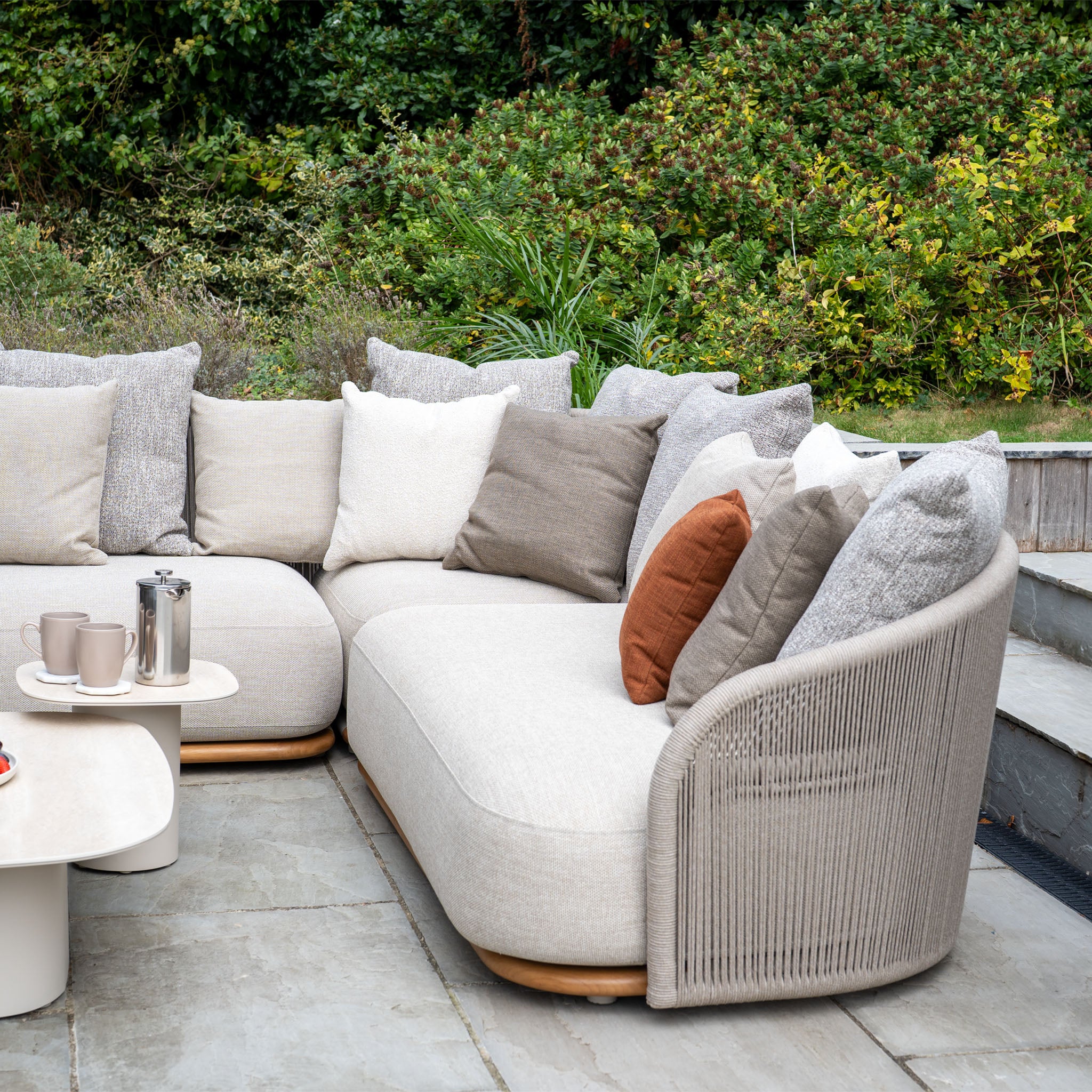 The Avola Corner Sofa Set in Fawn features modern, weather-resistant olefin cushions and a table with cups, perfect for outdoor spaces surrounded by lush greenery.