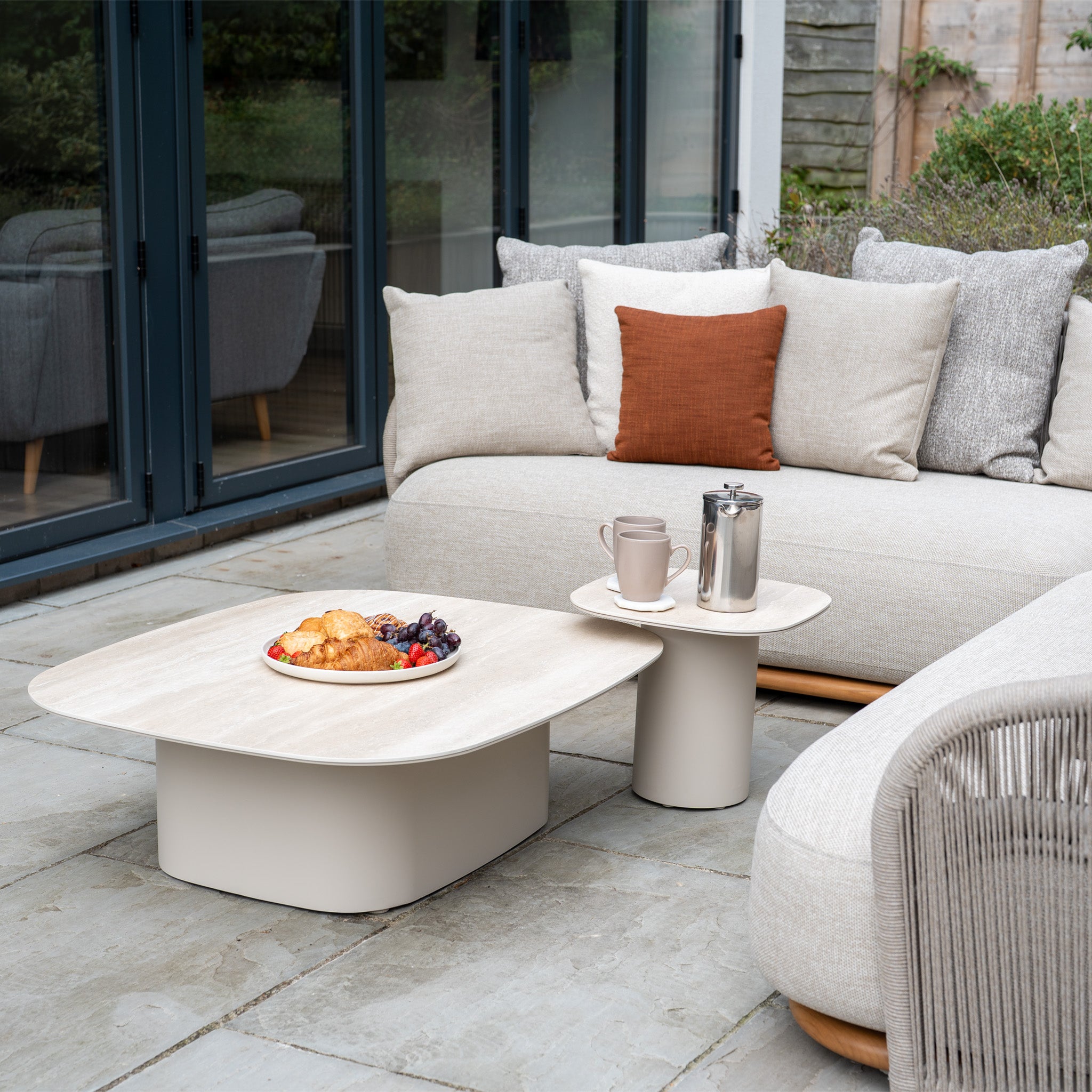 The Avola Corner Sofa Set in Fawn features weather-resistant beige cushions and a coffee table—perfect for stylish outdoor relaxation with pastries, fresh fruit, cups, and a French press.