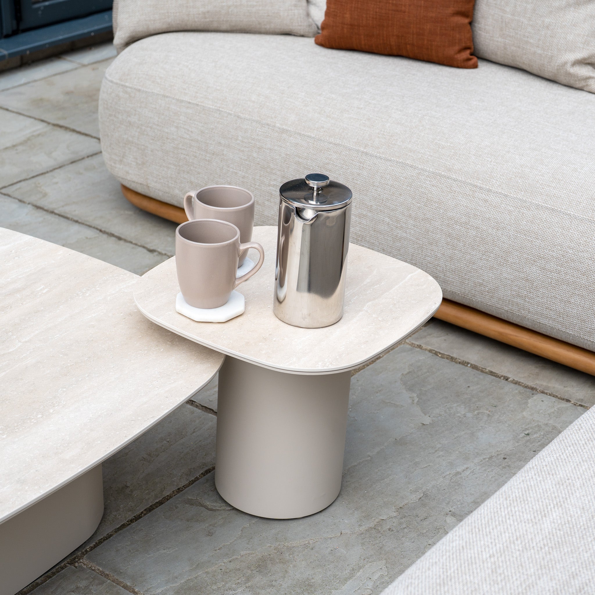 A small table with two mugs and a French press sits beside the Avola Corner Sofa Set in Fawn, a beige, weather-resistant outdoor garden sofa on a stone patio.